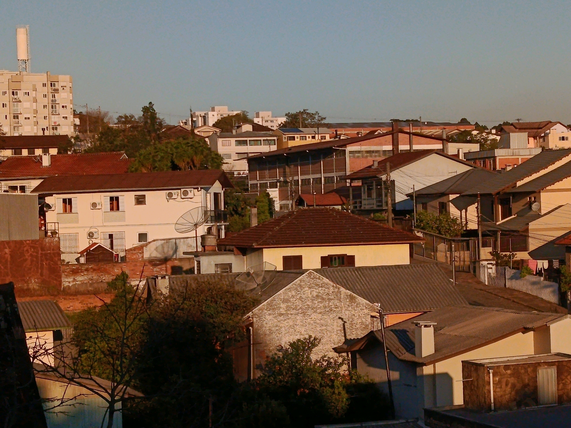 This is a daylight, eye-level shot of a dense urban neighborhood in Caxias Do Sul, Brazil, featuring a variety of residential buildings and a clear, pale blue sky. The buildings are primarily low to mid-rise, constructed with concrete, brick, and tile. Many exhibit signs of wear and tear, with peeling paint and weathered facades. Dominant colors include muted earth tones, whites, and reds.
In the background, a tall, white, cylindrical water tower stands out against the sky, likely serving the surrounding community. To its left, a multi-story apartment building with numerous balconies and windows is visible. The mid-ground is a tapestry of rooftops of varying materials and pitches, including corrugated metal and red tiles. Some houses have satellite dishes and air conditioning units visible on their exteriors. Green foliage from trees and bushes peeks through the gaps between buildings, suggesting a residential area with some vegetation.
The foreground is partially obscured by the shadows of nearby structures and dense trees. In the lower left, a dark, leafless tree is silhouetted against lighter surfaces. A small, white, single-story structure with a dark roof is partially visible in the lower left quadrant. The overall impression is of a lived-in, somewhat aged urban environment. No people are visible in the scene, and no specific actions or interactions are depicted. The focus is purely on the built environment and the sky.