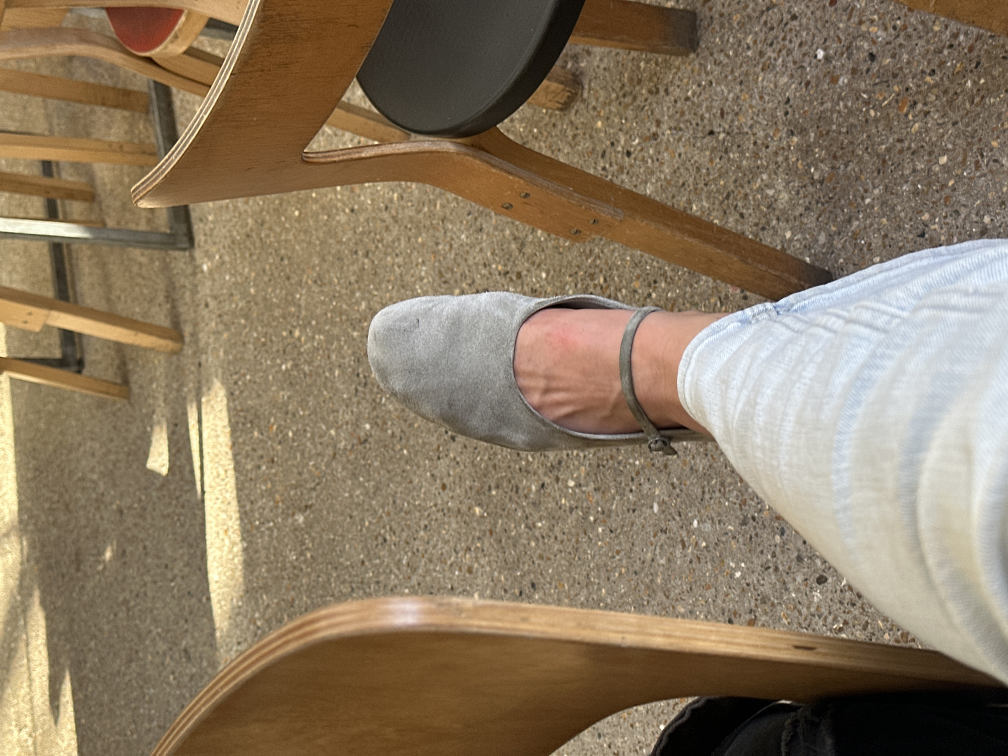 The image captures a close-up, angled view of a person's lower leg and foot, seated in what appears to be a public or commercial setting in London, United Kingdom. The person is wearing light-colored pants, possibly light blue or off-white with a subtle striped texture, and a grey flat shoe made of a suede-like material. The shoe features a thin strap across the instep.
Surrounding the leg are various pieces of wooden furniture, including chairs or stools, some with visible metal elements. One chair in the mid-ground has a distinct dark, circular seat. The wooden components are a light to medium brown, showing some wear and visible grain.
The floor is a textured, speckled surface, resembling light grey or beige concrete or terrazzo with small, multi-colored aggregate chips. Bright sunlight streams into the scene, casting sharp, elongated shadows across the floor and highlighting parts of the furniture, suggesting it is daytime, likely morning or afternoon. There is no visible text in the image, and no specific emotions are discernible. The person's activity is simply being seated.