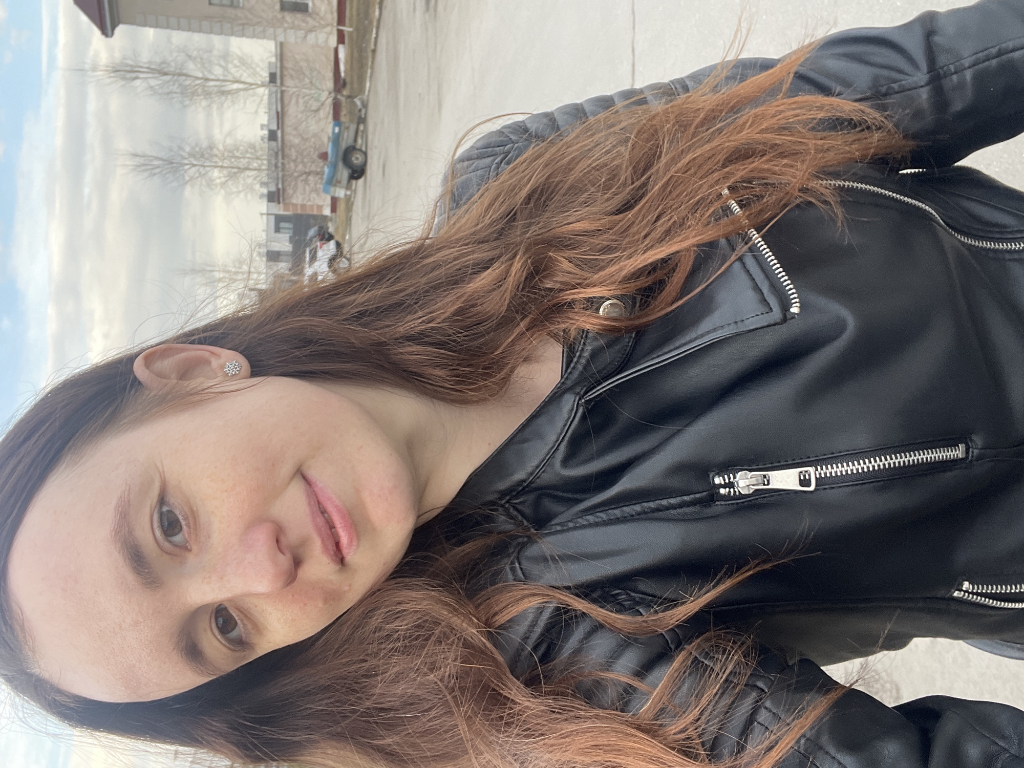 The image shows a woman with long, wavy auburn hair and fair skin, wearing a black leather jacket. She is oriented diagonally in the frame, facing the viewer with a slight smile. The background shows a cityscape with a building and trees in the distance, under a partly cloudy sky. Based on geographic information, the scene is located in the city of Подстёпки, Russia.