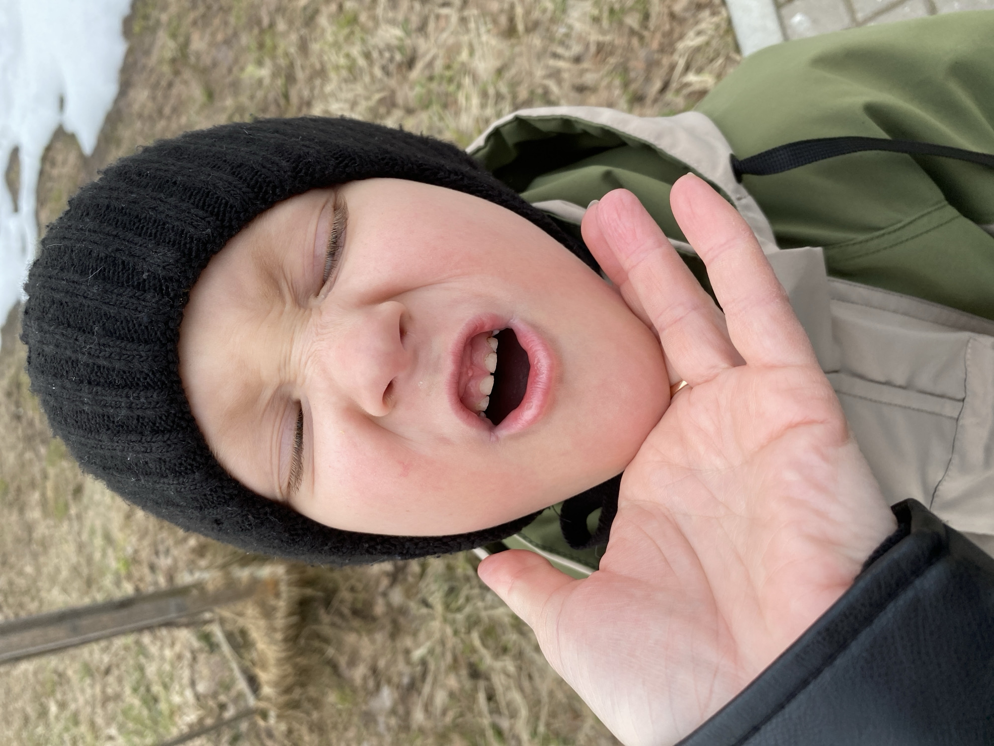 A young person with closed eyes is captured in a close-up shot. They have a black knit cap on their head and are wearing a jacket in shades of green and beige. The person's mouth is open, revealing their teeth. A hand with a ring on one finger is touching the person's face. The background includes a mix of grass, snow, and a sidewalk or paved area. The image appears to have been taken outdoors in Noviy, Russia.