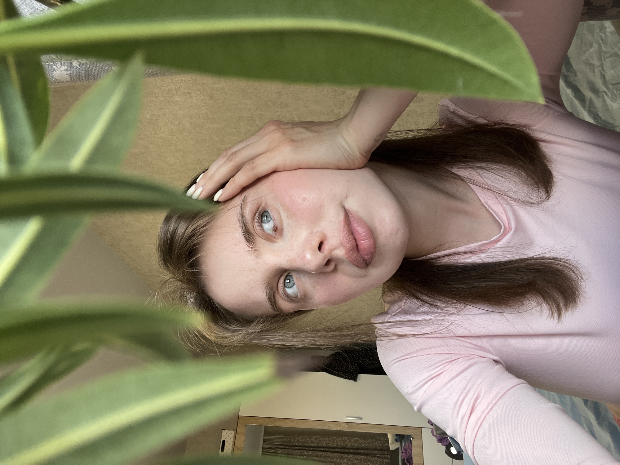 A woman with fair skin, light blue eyes, and medium brown hair is seen in a selfie. Her left hand is resting on her forehead. She is wearing a long-sleeved pink shirt. Large green leaves of a plant are prominently featured in the foreground, partially obscuring the view. The background includes a light brown surface, possibly a wall or tabletop, and a glimpse of furniture. The location is likely indoors. There is no verifiable information to confirm this photo was taken in Chaikovsky, Russia.