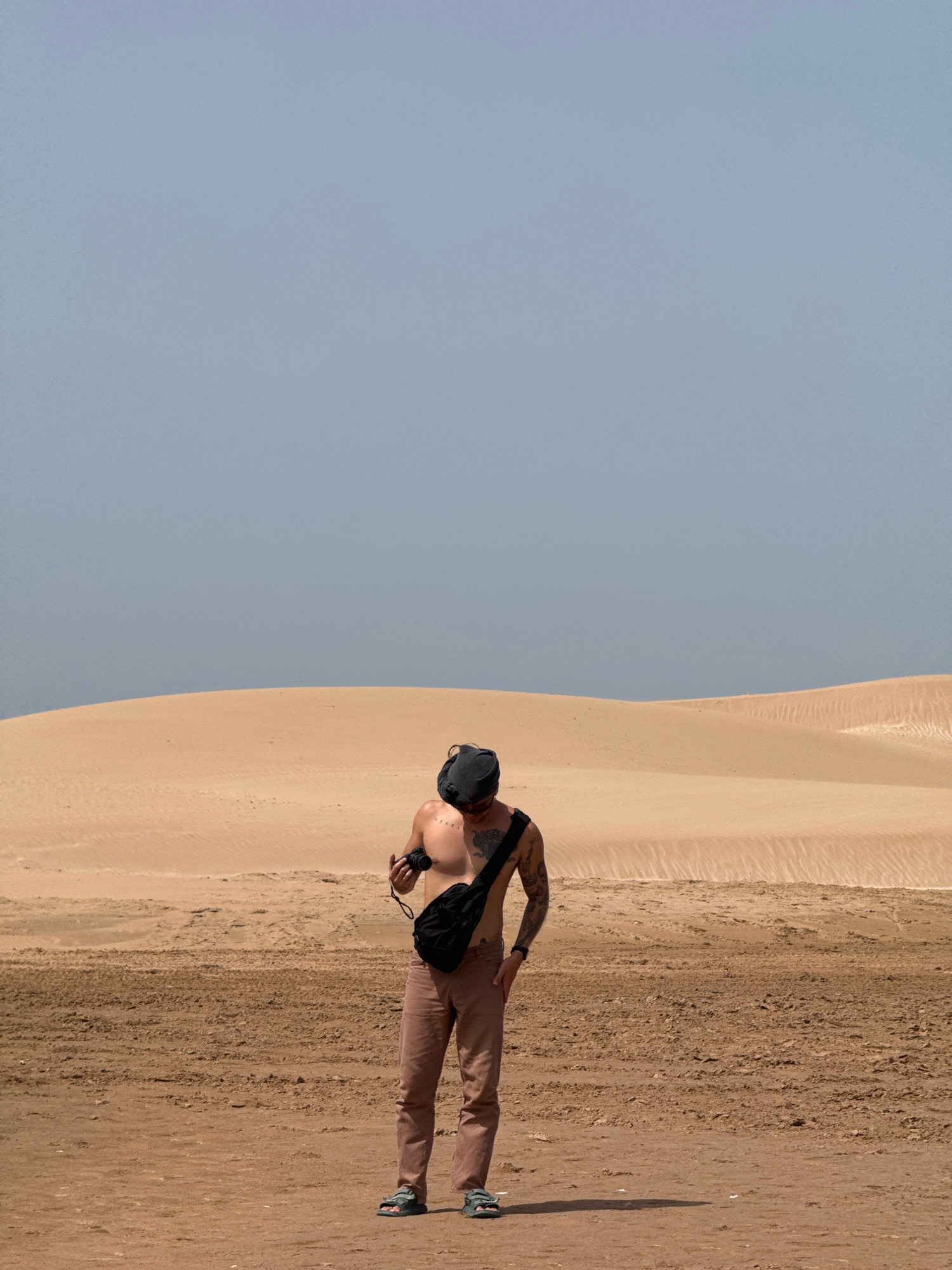 In the city of Essaouira, Morocco, a shirtless man wearing a black cap and brown trousers stands in a desert landscape. He has a black fanny pack across his chest and is holding a camera in his right hand. The background consists of sand dunes under a clear, pale blue sky.