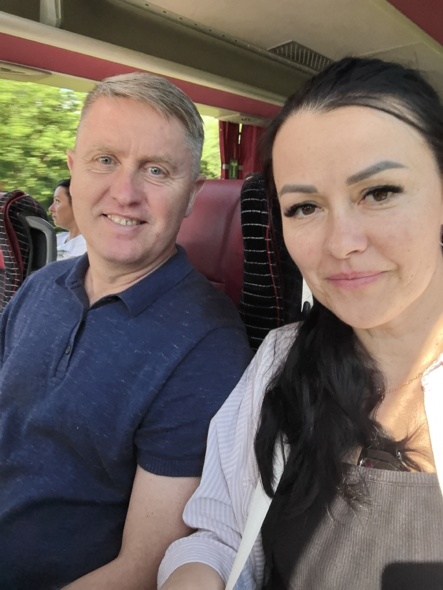 The image is a selfie of a couple on a bus. The man in the foreground on the left is smiling, wearing a navy blue polo shirt. His hair is short and graying, and he has blue eyes. Beside him on the right is a woman with long, dark hair. She is also smiling and wearing a light-colored striped shirt and a gray tank top underneath. Her eyes are brown, and she has noticeable makeup, including false eyelashes.
In the background, out of focus, other passengers can be seen on the bus. The windows of the bus show a blur of green trees, suggesting the bus is moving through a scenic, possibly rural area. The interior of the bus has red seats and what appear to be window shades. The lighting suggests it is daytime, likely late afternoon or early evening due to the warm, angled sunlight. There are no visible text elements in the image. The overall mood is relaxed and happy, consistent with a couple enjoying a trip together.