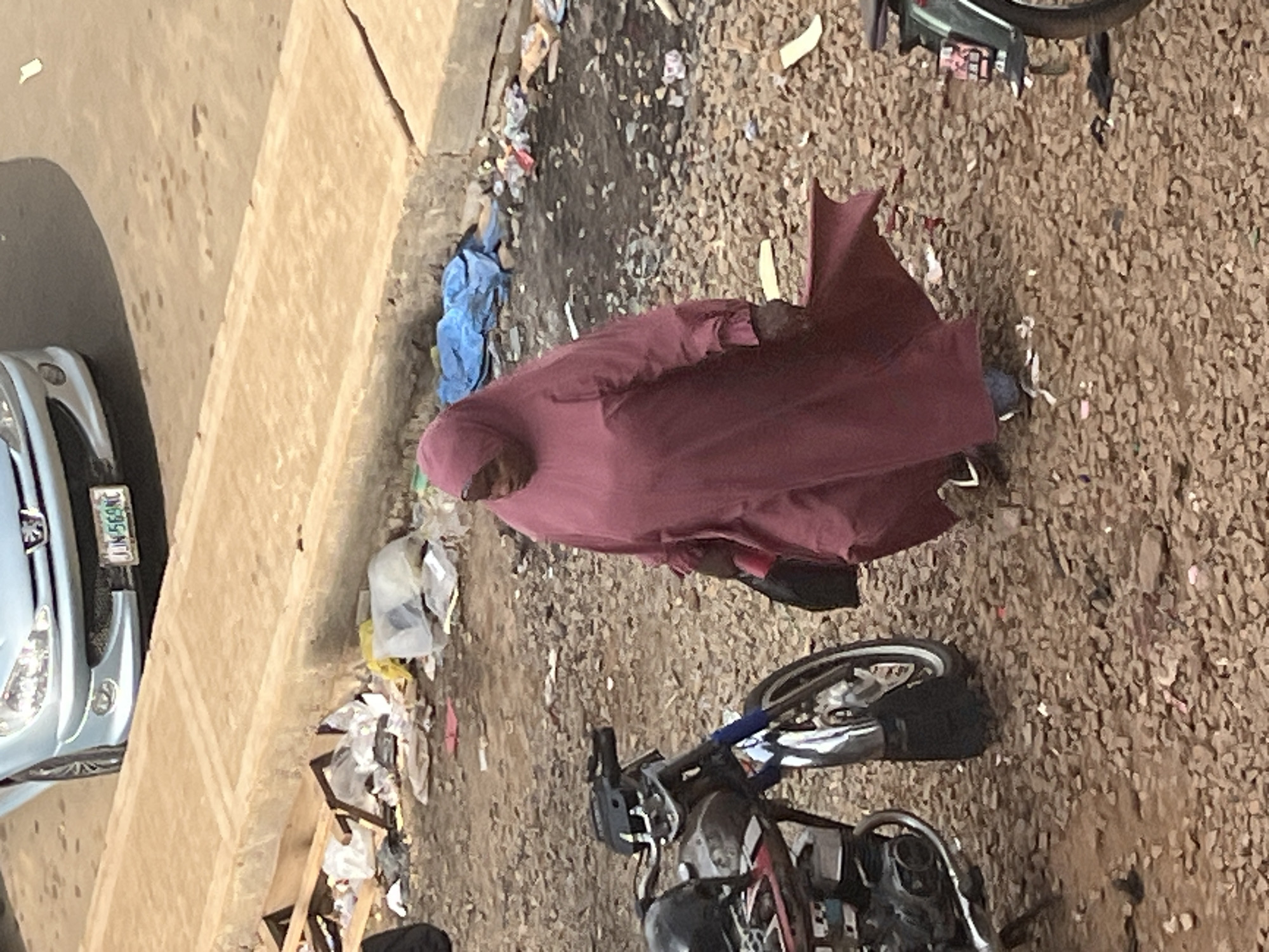 In Jos, Nigeria, a person wearing a maroon hijab and abaya stands in a lot filled with debris. The ground is covered in small stones and scattered trash, including plastic bags and other discarded items. To the left, the front of a light blue car is visible, with its license plate reading "NJN 569NC". In the foreground and slightly to the right, a motorcycle is parked. The scene appears to be an outdoor area, possibly a street or lot in a less-maintained part of the city. The lighting suggests it might be daytime, with shadows indicating sunlight. The presence of a car, motorcycle, and a person in traditional attire provides context about the location. There is no clear indication of the specific activity or event taking place, other than the person standing amidst the surroundings. The overall impression is one of everyday life in a bustling environment with visible signs of unmanaged waste.