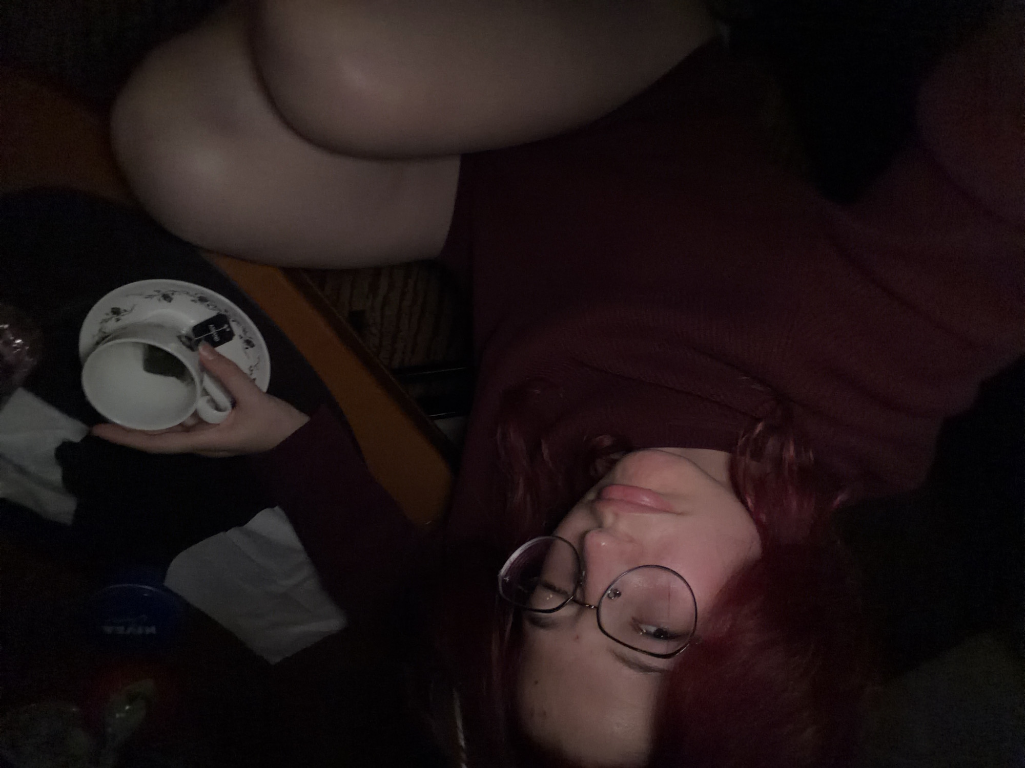 The image is a first-person, low-light selfie taken indoors. A person with reddish hair and glasses is looking at the camera with a pouty expression. They are wearing a dark reddish-brown sweater. To their right, on a dark surface, is a teacup and saucer with a teabag in the cup. In the background, a blue NIVEA container is visible. The environment appears to be a room in a residential setting. There are no identifiable landmarks or features that would indicate a specific location within Chelyabinsk, Russia.
