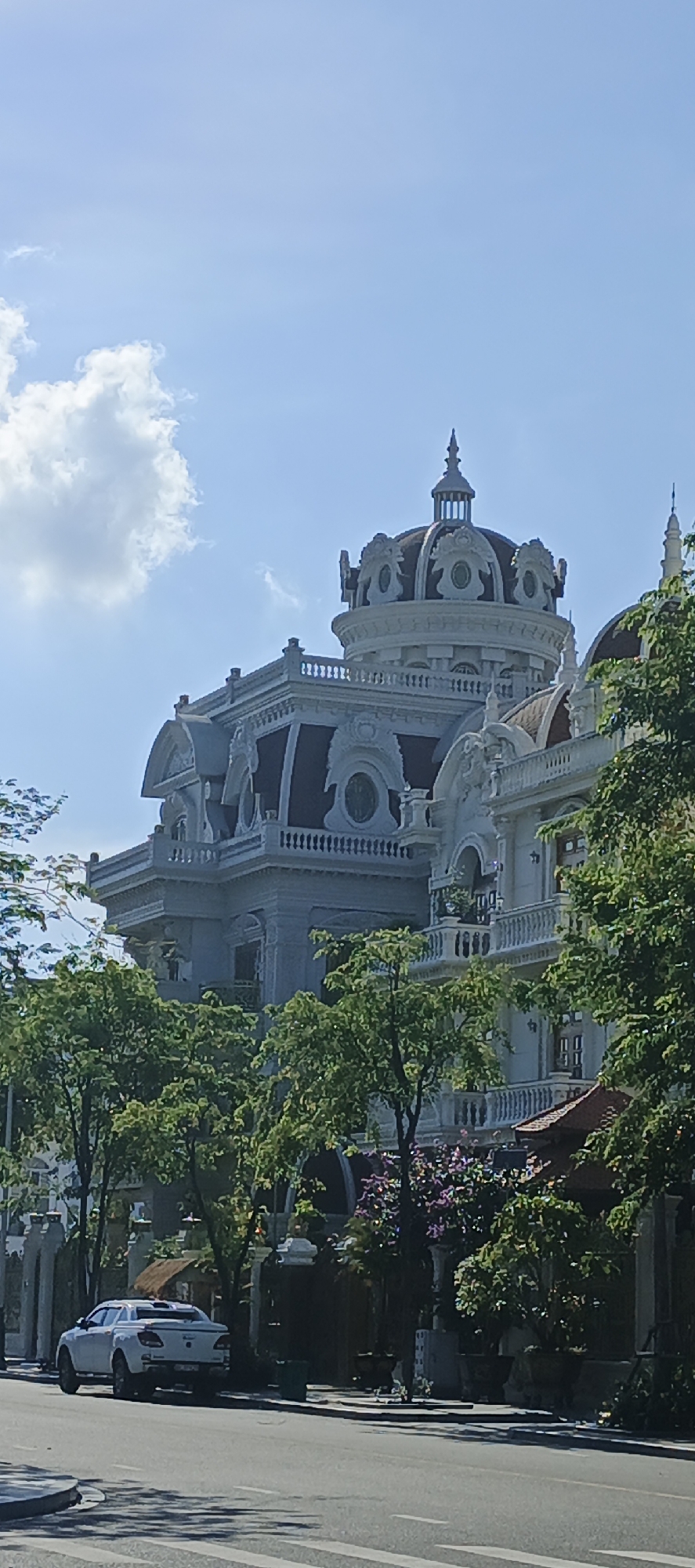 In an urban setting under a clear blue sky with scattered clouds, a white pickup truck is parked on the side of a paved road. The truck is facing left, with its rear toward the viewer. Behind the truck, a large, ornate building with white facade and dark-tiled roofs dominates the scene. The building features classical architectural elements such as domes, balconies, and detailed carvings. Lush green trees and purple flowering plants are in the foreground, partially obscuring the lower levels of the building. The street has crosswalk markings.