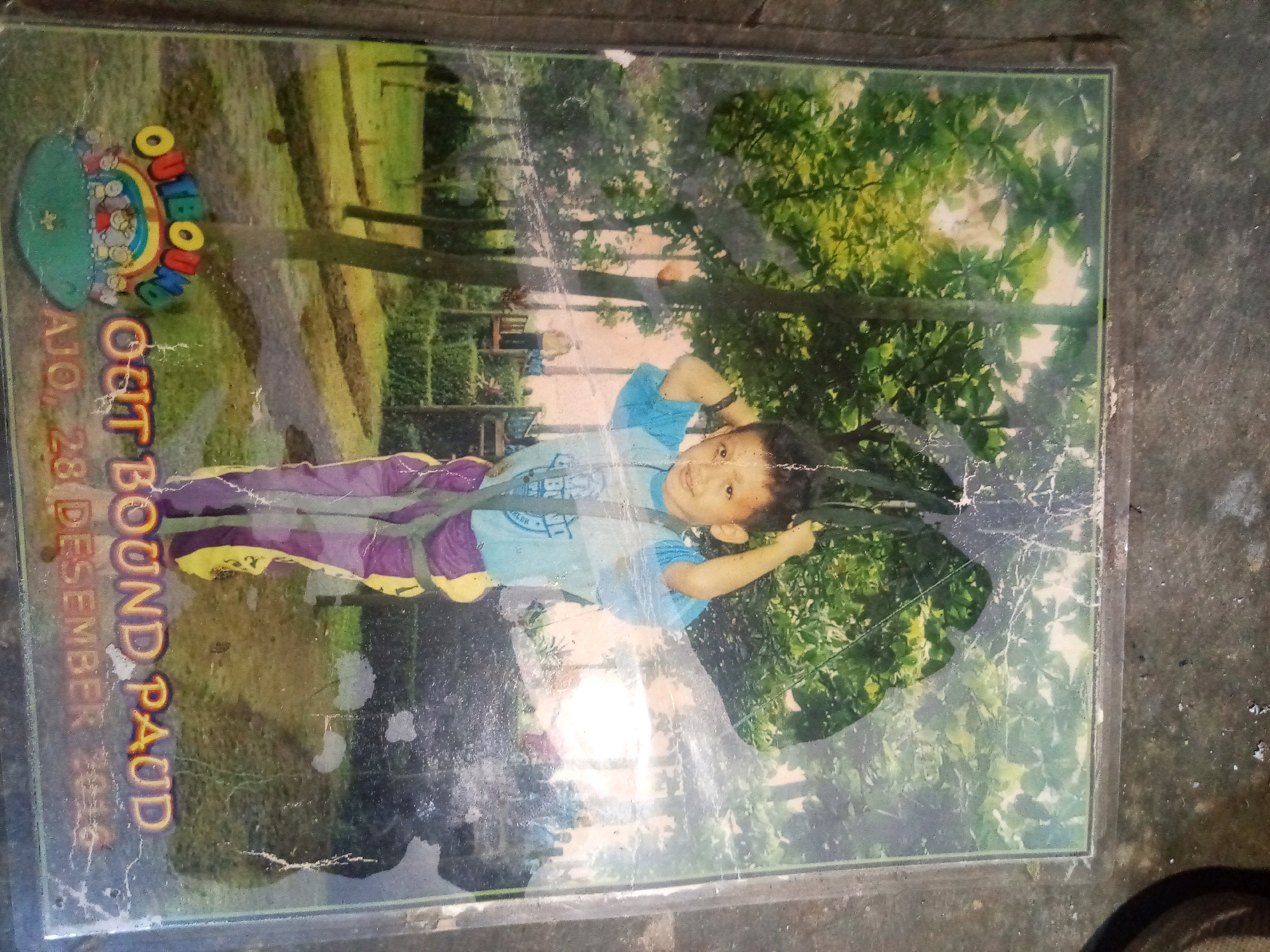 The image shows a photograph of a young boy lying on his back outdoors, with his head propped up on his hand. He is wearing a light blue t-shirt and purple pants. The photograph is oriented vertically. Text on the left side of the photograph reads "OUT BOUND PAUD" and "AJO, 28 DESEMBER 2015". The background of the photograph appears to be a park or grassy area with trees and sunlight filtering through the leaves. The photograph itself is framed and slightly scratched. The overall scene is a captured moment of childhood activity.