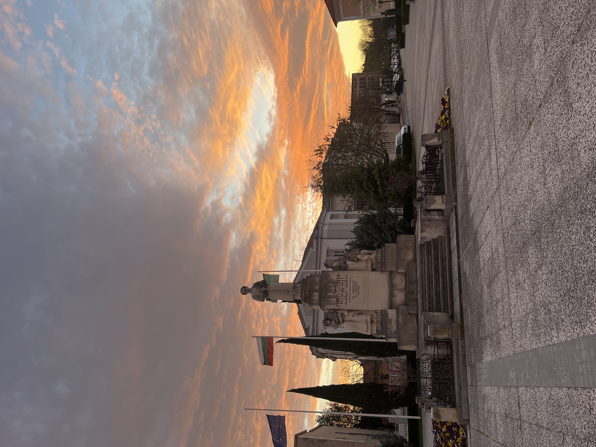 The image depicts a monument with flags in Nova Zagora, Bulgaria, set against a vibrant sunset. The monument is made of stone and features statues of people, including a figure at the top. The Bulgarian flag and the European Union flag are prominently displayed. The sky is filled with clouds tinged with orange and pink hues from the setting sun. The foreground shows a paved area and landscaping around the monument. Buildings and trees are visible in the background.