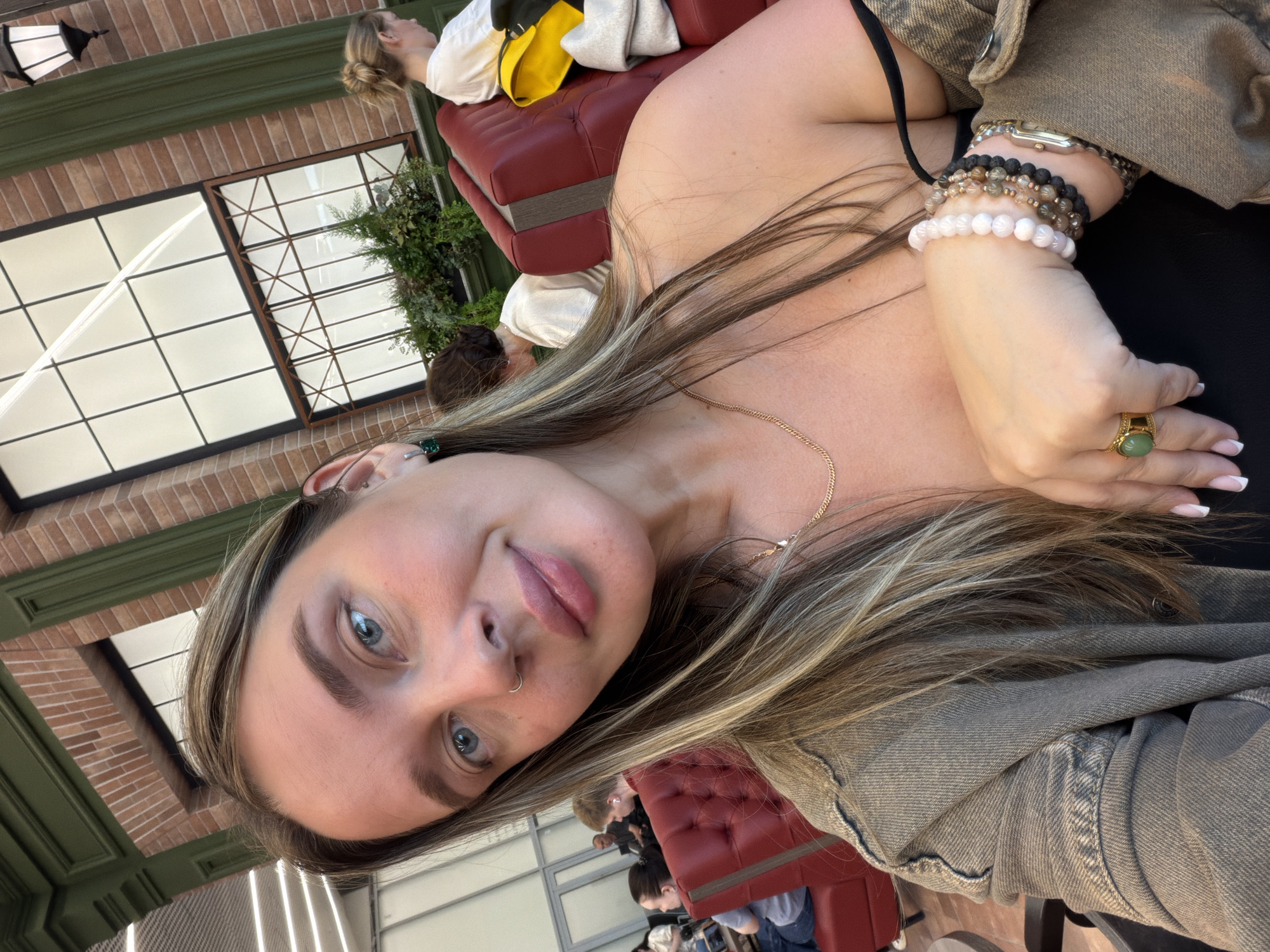 The image shows a young woman taking a selfie in a cafe. She has blonde hair, blue eyes, and is wearing a denim jacket. She is also wearing several bracelets and a ring. The cafe appears to be modern, with red leather booths and large windows. In the background, other patrons can be seen seated at tables. The woman is smiling and looking at the camera. The scene is well-lit and the focus is on the woman's face. It is not possible to determine the location within the city of Izhevsk, Russia based on the provided image.