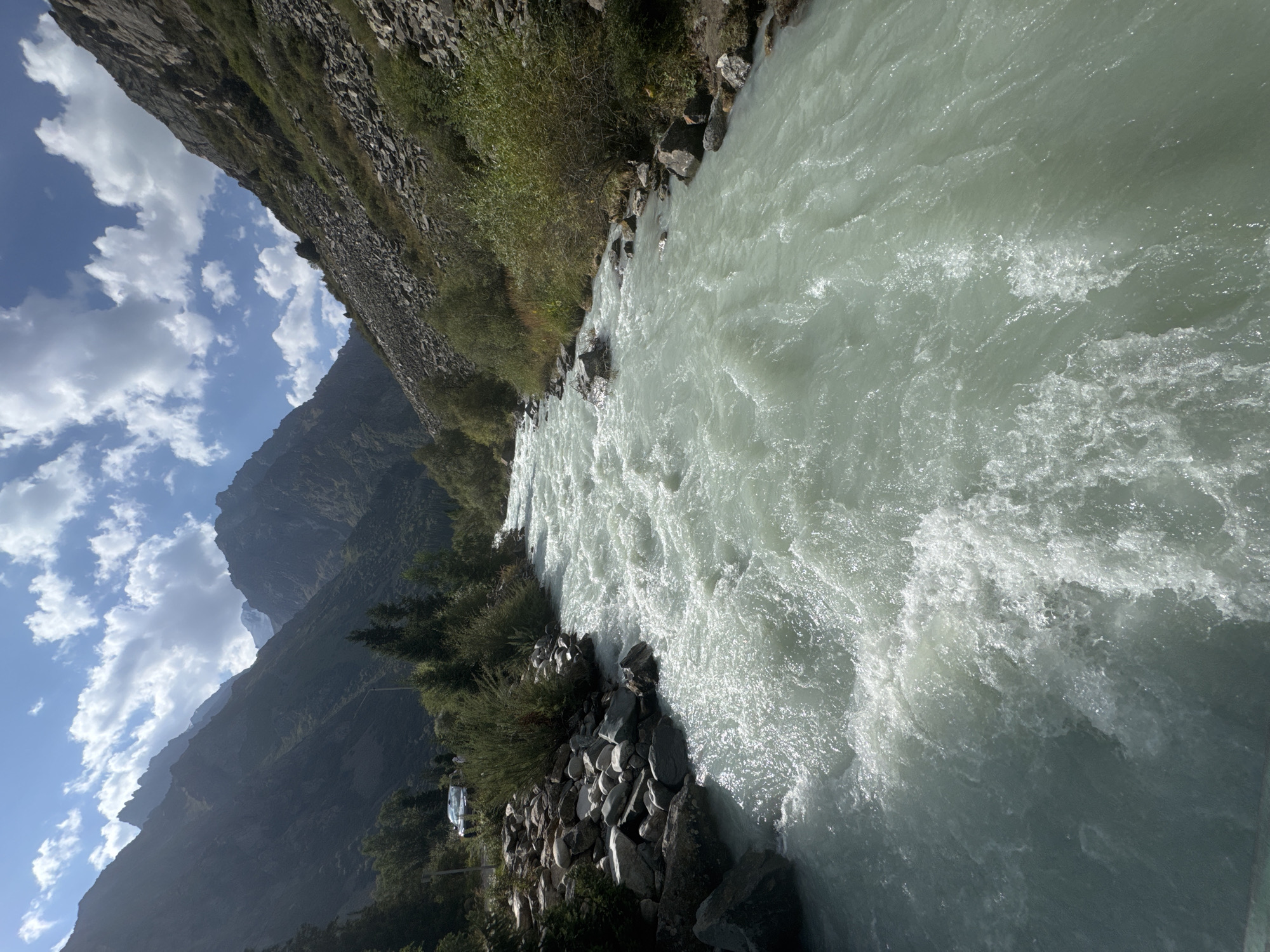 The image depicts a wide, turbulent river flowing through a mountainous valley. The water is a milky, light green color, indicative of glacial melt. The riverbanks are lined with rocks and vegetation, with steeper, rocky slopes rising on either side of the valley. In the background, jagged mountain peaks are visible under a bright blue sky dotted with white, fluffy clouds. On the left side of the river, near the bank, there is a small area with a car and some sparse trees. The overall scene conveys a sense of wild, natural beauty. This is likely a scene from Ala Archa National Park, near Bishkek, Kyrgyzstan, a popular destination known for its glaciers, rivers, and mountains.