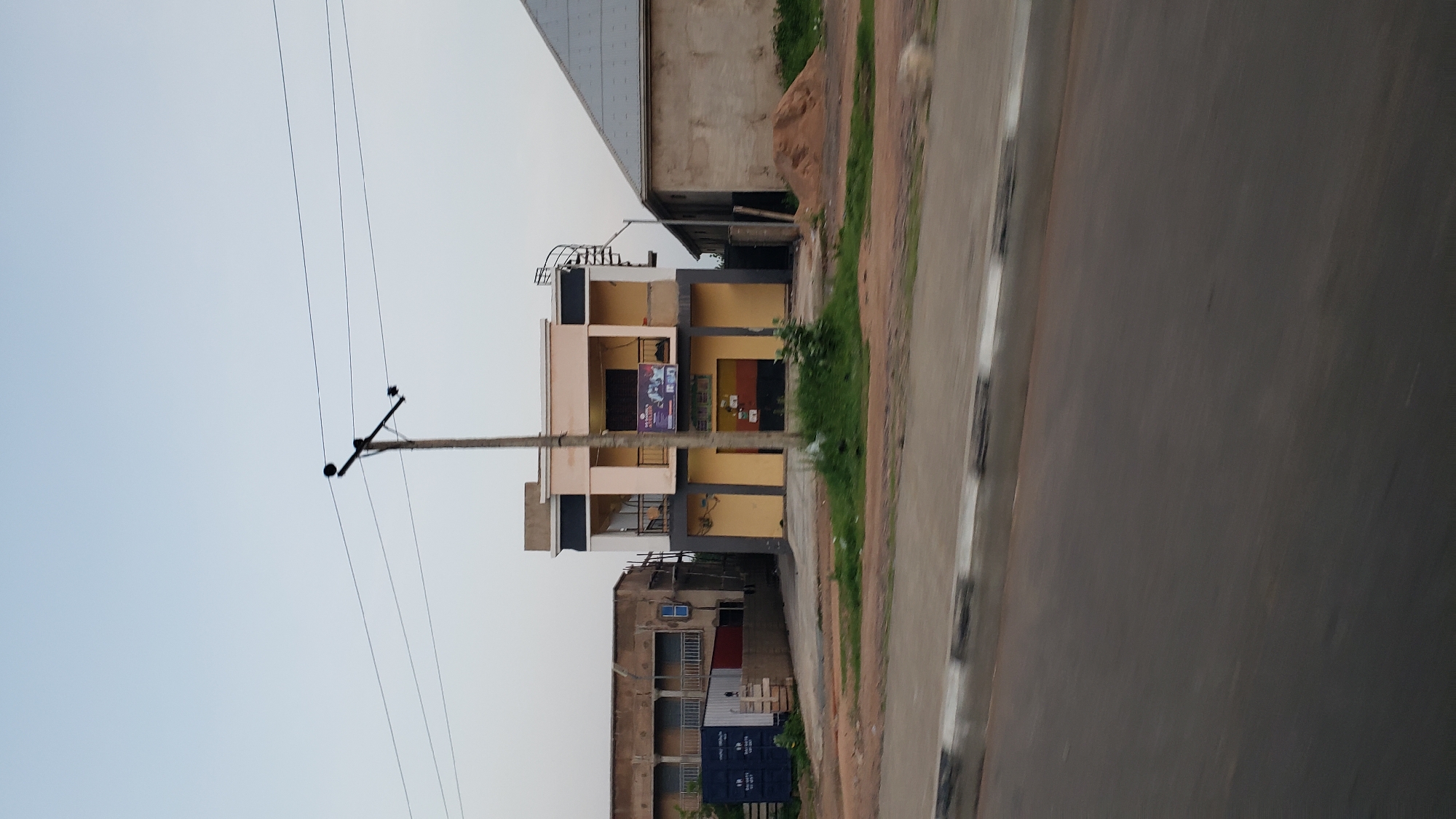 The image depicts a street scene in Jalingo, Nigeria. The focal point is a two-story building with a light yellow and beige facade, featuring a dark grey accent band. The ground floor has multiple shop fronts, with one displaying a sign that reads "MUMU AYELID". Above the shops, a balcony with a black railing is visible. To the right of this building, a spiral staircase is attached to the exterior, leading to the upper level. Adjacent to this building, on its right, is a portion of another structure with a grey, angled roof and concrete walls. To the left of the main building, a multi-story building with balconies and what appears to be ongoing construction or renovation is present, along with a large blue shipping container. A utility pole with overhead wires stands prominently in front of the two-story building. The street in the foreground is paved and shows a grey curb with white markings. The overall environment appears to be a commercial area with residential or mixed-use buildings. The sky is a pale, overcast blue, suggesting it might be early morning or late afternoon. No people are clearly visible in the image, and no specific actions or interactions are depicted.