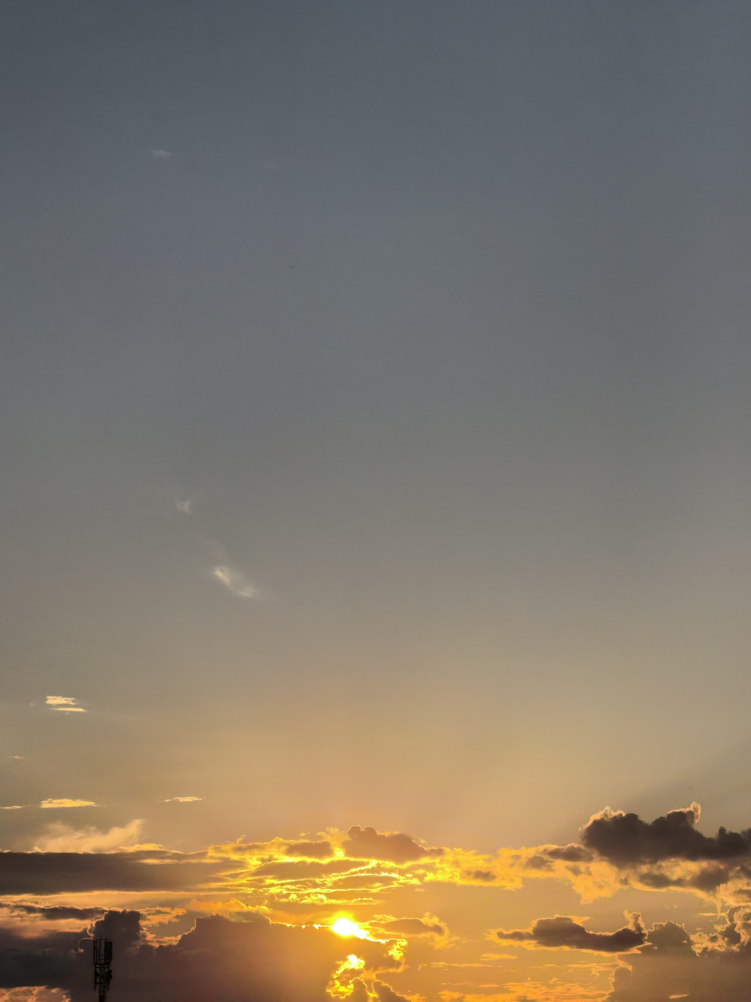 An outdoor, vertical, full-frame shot captures a dramatic sunset over the city of Unknown, Unknown. The sky transitions from a muted gray at the top to warm oranges and yellows near the horizon. Scattered clouds are present, with some illuminated by the setting sun, creating a vibrant, fiery effect. A tall, slender cell tower is visible in the lower left portion of the image, silhouetted against the bright sky. The sun itself is a brilliant, intense yellow sphere, partially obscured by clouds and casting radiant rays. The overall mood is one of natural beauty and the end of the day.