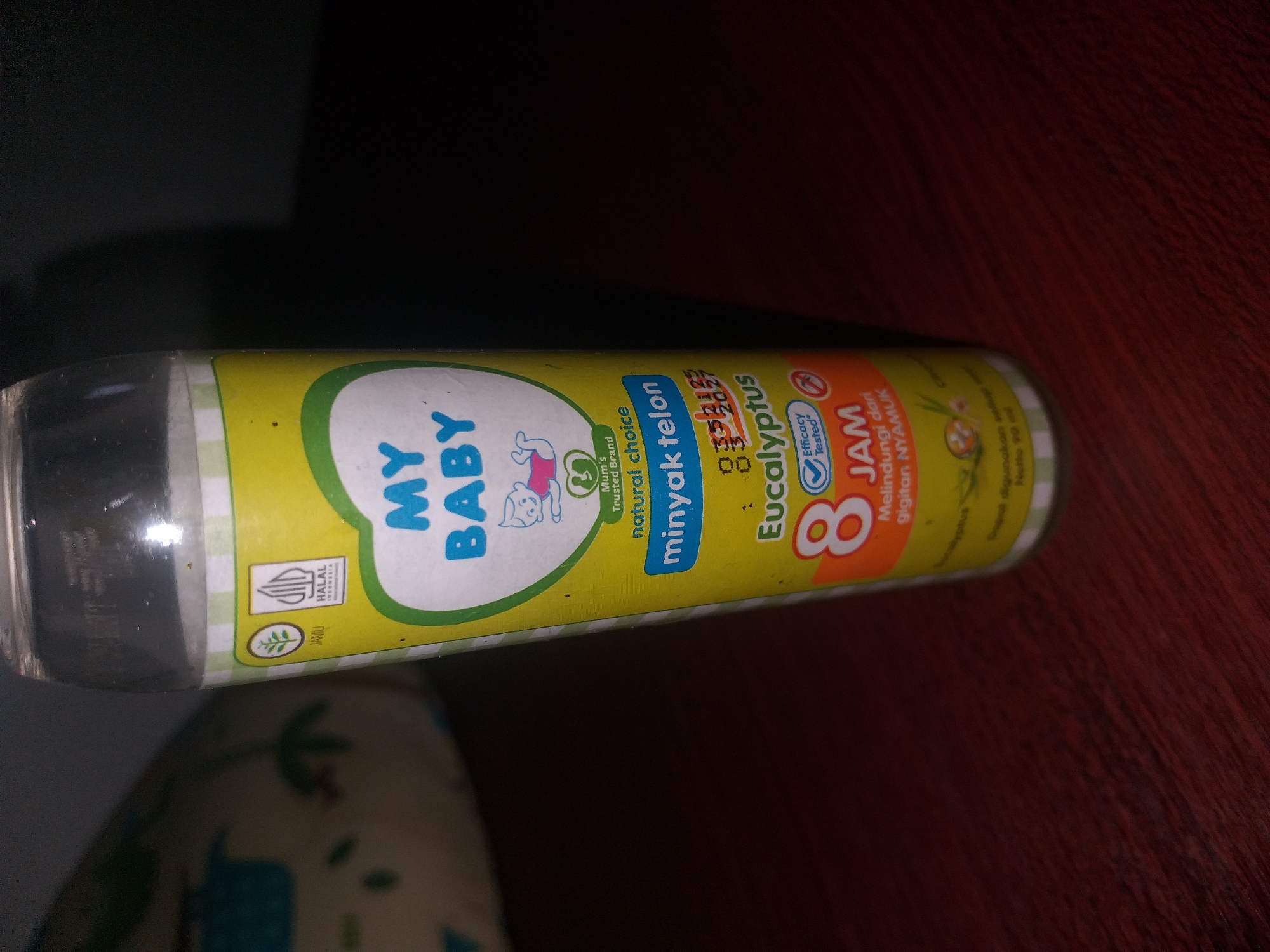 A close-up, vertical, eye-level shot shows a bottle of My Baby brand eucalyptus oil, with a yellow label, on a dark wooden surface. The bottle is partially obscured by darkness. The label features a cartoon of a baby, text that reads "MY BABY," "Minyak telon," and "Eucalyptus," as well as "8 JAM" indicating 8 hours of protection. It also has a "HALAL" logo. The background shows a blurred, light-colored fabric object with green leaf patterns. There is no indication of the specific location within Air Jamban, Indonesia.