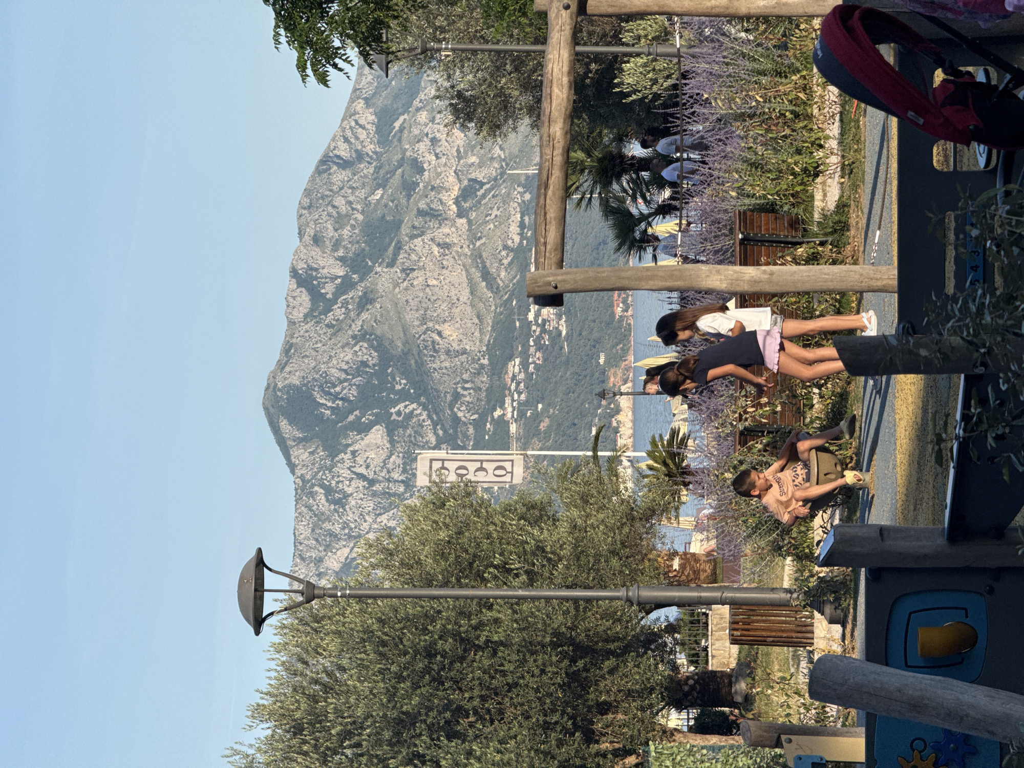 The image, originally rotated 90 degrees clockwise, depicts a vibrant daylight scene of children playing in a park or playground situated along a mountainous coast, characteristic of Budva, Montenegro.
In the foreground, several children are visible. Two girls stand near a wooden playground structure; one wears a white t-shirt and light shorts, while the other is in a dark top and pink shorts. To their right, a boy in a light t-shirt with the word "Strengt" (likely "Strength") and patterned shorts sits on a spring-rocker. Parts of a blue and yellow play structure are also visible at the bottom right. The playground area features green foliage, including olive trees and flowering purple lavender bushes. A black lamppost stands on the left.
The midground and background reveal a dramatic landscape. A large, rugged grey mountain dominates the left side of the frame. Below it, the calm blue sea stretches out, with numerous buildings of a coastal town nestled along the shoreline and what appear to be beach umbrellas. A white rectangular sign with four black circular symbols is visible in the distance, though its specific function is unclear. The sky is a clear, bright blue, indicating sunny weather. The children appear engaged in play, contributing to a lively and picturesque atmosphere.