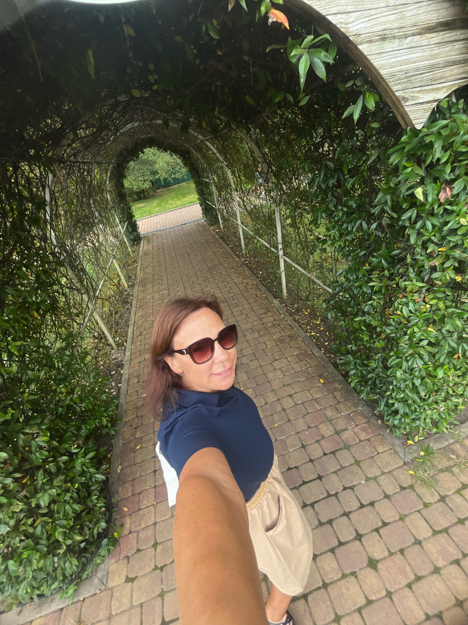 The image shows a woman taking a selfie in a green leafy tunnel. She is wearing sunglasses, a dark blue shirt, and a beige skirt. The tunnel is made of arched trellises covered with vines and leaves, with a paved walkway underneath. The walkway is made of brown and gray cobblestones. In the background, there is a glimpse of a green lawn and trees, suggesting an outdoor location such as a park or garden. The overall lighting is somewhat dim, indicative of a shaded area. The woman is looking at the camera and smiling slightly. The environment appears to be well-maintained and natural. There is no indication of the specific location within the city of Sochi, Russia.