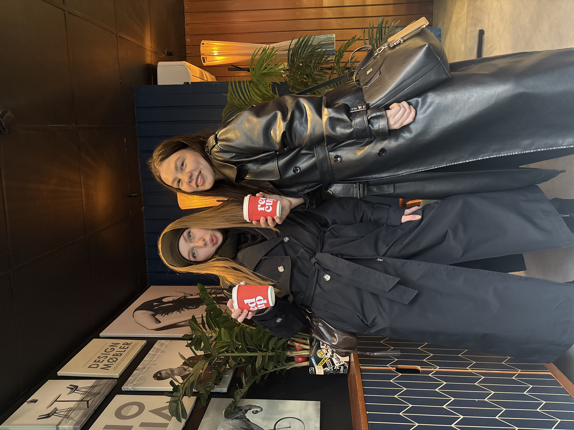 The image shows two young women posing in a modern indoor setting, likely a cafe or restaurant in Perm, Russia. Both women are wearing dark trench coats, suggesting a fashionable outing.
The woman positioned on the left has long, dark hair and a bright smile directed at the camera. She wears a black faux leather trench coat, with a black handbag draped over her arm. She holds a red disposable cup with the text "RED CUP" clearly visible in white.
The woman on the right has long, lighter hair, partially covered by a dark beanie. She looks towards the camera with a playful pout or smirk. She holds two identical red disposable cups, each also bearing the "RED CUP" text in white. She is dressed in a black trench coat with a belted waist.
The background features contemporary interior design. To the left, a collection of framed wall art or posters is displayed. One poster explicitly reads "DESIGN MØBLER" (Danish for "Design Furniture"), and another shows the partial text "HO." Other posters depict abstract figures, an elephant, and a bicycle. Behind the women, dark wood paneling, a dark blue textured wall or cabinet, and concrete-like walls are visible. Green potted plants are strategically placed, and warm, ambient lighting emanates from decorative lamps with textured shades. A table or shelf in the foreground exhibits a distinctive black and gold geometric pattern. The scene conveys a casual and stylish atmosphere.
