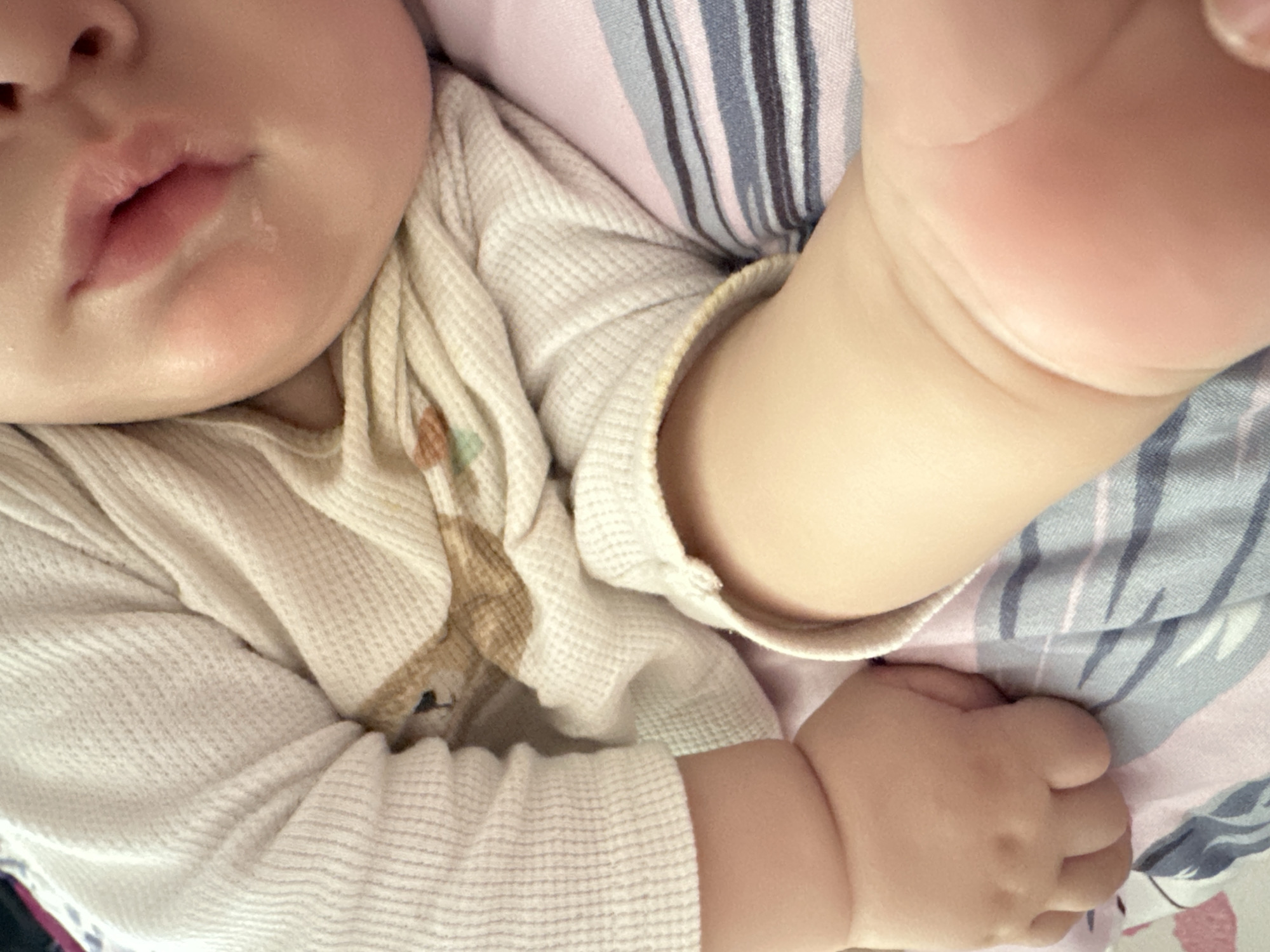 The image shows a close-up of a baby lying down. The baby is wearing a white thermal shirt with a brown and green design on the chest. A small amount of drool is visible on the baby's lip. The baby's left hand is visible, and the right arm is partially in view. The baby is lying on a surface with a pink, blue, and white striped pattern.