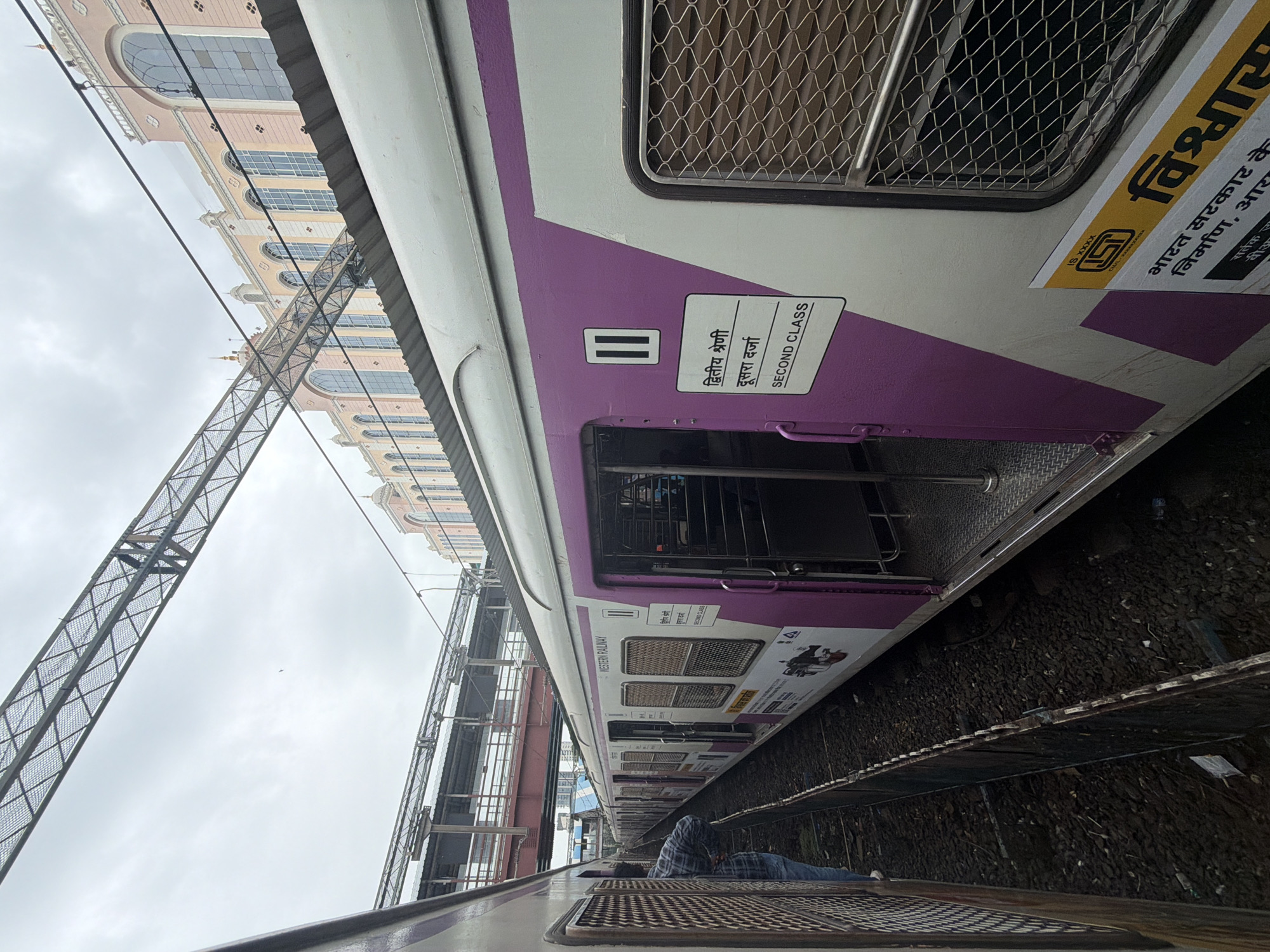 This is a photograph of a Mumbai suburban railway train at a station, likely on the Western Line, given the "WESTERN RAILWAY" visible on the train car. The train is parked on tracks, with a platform visible to the left. The train car is primarily white with a distinctive purple diagonal stripe pattern. A sign on the side of the car indicates "SECOND CLASS" in English and Hindi. Several doors on the train are open, revealing seating inside. The background shows a railway bridge and the facade of a pink and cream colored building with arched windows. The sky is overcast. A person in a blue and white checkered shirt and jeans is standing near one of the open train doors, looking away from the camera. The ground around the tracks is covered with ballast and debris.