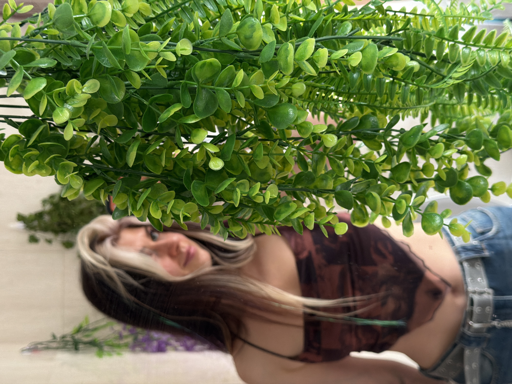 The image presents a unique composition, appearing to be a reflection of a person viewed through or beneath a dense layer of artificial greenery. The upper portion of the frame is dominated by a variety of vibrant green artificial plants with small, glossy leaves and thin stems, some rounded and others more elongated, creating a lush, textured foreground that is sharply in focus.
Beneath this foliage, the blurred reflection of a person, likely female, is visible, positioned diagonally across the frame. The person has long, brown hair with distinct blonde highlights. Their face is partially obscured and out of focus, showing soft lips and part of an eye. They are wearing a dark, possibly reddish-brown or earthy-toned patterned top with thin straps, paired with blue denim jeans and a prominent silver-buckled belt.
The activity suggests the person is posing for a photo, likely taken into a mirror or other reflective surface. In the background of the reflection, hints of other elements, such as a blurred bundle of purple flowers and more greenery, can be seen on the surface. The lighting is soft and even, typical of an indoor setting. There is no visible text in the image. The overall impression is casual and artistic due to the layered depth and the use of reflection. The provided context for this media file is Budva, Montenegro.