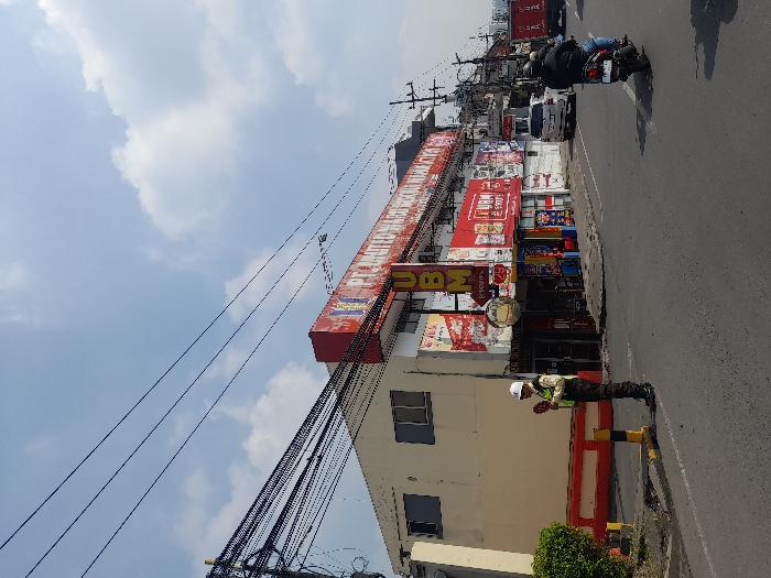 The scene is in front of a large, two-story building with multiple shops and businesses. The building has a large, red and white sign with the words "PT. UNITOMO BERSAMA" visible, which translates to "PT. Unitomo Together". There are also signs with the word "UBM" (possibly an abbreviation for a store name) in bold and other, smaller signs, along with several windows, some open and some closed. A man in a white and blue uniform stands outside, holding a red object. There is a red, yellow, and black barrier on the ground in front of him. The street is paved asphalt with a concrete sidewalk. In the background, a white van is parked to the left, with a red motorcycle to the right. There is a man riding the motorcycle. The sky is blue with white clouds. Power lines run across the sky from the left to the right side of the scene. The location is on a street in the city of Kecamatan Waru, Indonesia, based on the coordinates -7.354 latitude and 112.729 longitude.