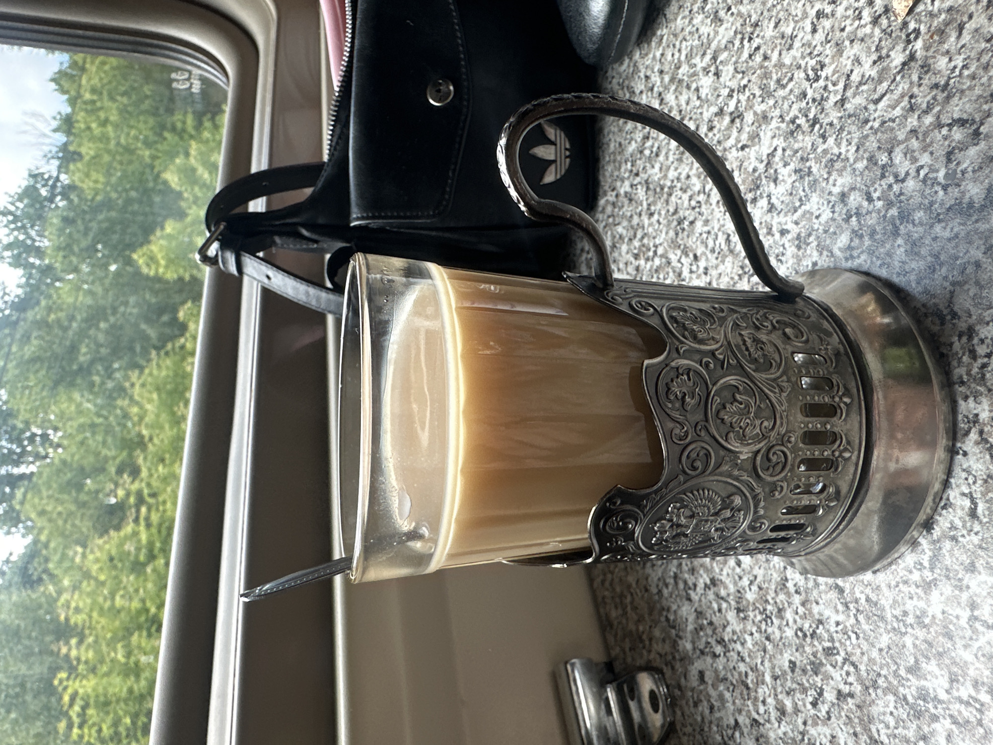 The image captures a tranquil moment within what appears to be a train car, seen from a passenger's perspective. The central focus is a clear glass filled with a light, milky beverage, likely coffee or tea with milk, held firmly in an ornate, silver-colored *podstakannik* (traditional Russian tea glass holder). A small spoon rests inside the glass. The intricate design of the *podstakannik* features swirling floral patterns and a central emblem.
Behind the glass, a black bag, possibly a handbag or small satchel, is visible. A prominent silver Adidas trefoil logo is embossed on the bag. The surface beneath the glass and bag is a speckled grey material, resembling granite or a similar durable countertop.
Through the window to the left, a vibrant green landscape with numerous trees under a bright sky suggests a sunny daytime journey, likely during spring or summer. The window frame and the general interior aesthetic are consistent with a train compartment.
The scene evokes a sense of calm and travel, characteristic of long-distance train journeys, where such glass holders are common, especially in Russia, aligning with the provided location context of Куркачи, Russia. No people are visible, but the presence of personal items implies a passenger enjoying the journey.