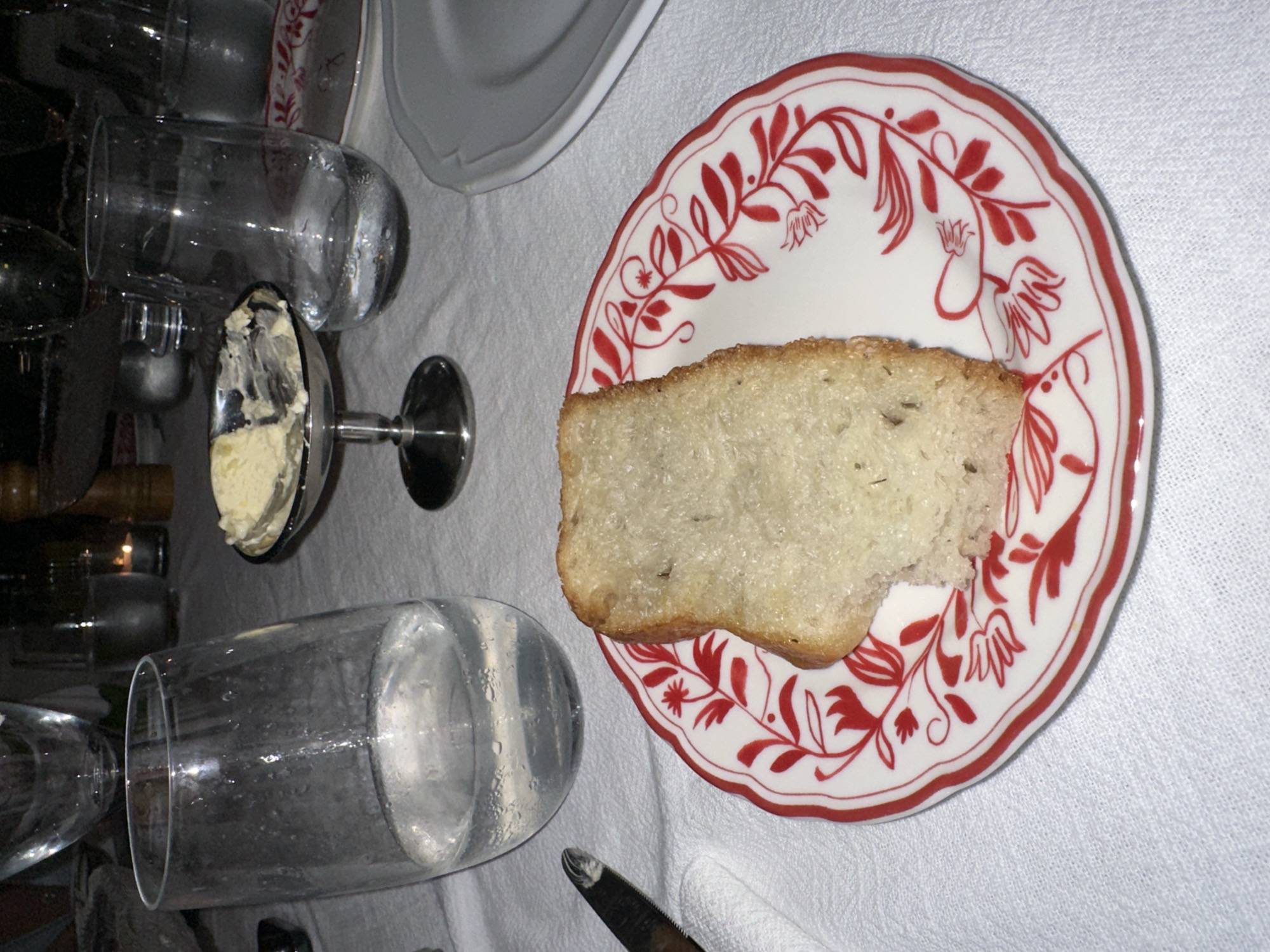 The rotated image captures a dining table setting, likely indoors. A white tablecloth serves as the backdrop for various meal accompaniments. In the center, a slice of light-colored, rustic bread rests on a white ceramic plate adorned with a distinctive red floral and vine pattern along its scalloped rim.
Several clear drinking glasses are visible, with one large glass in the foreground exhibiting condensation, indicating a cold beverage has been served or is intended. To the left, a silver, footed dish contains a creamy spread, likely butter, with a spoon partially submerged within it. A knife with a dark handle and silver blade is partially visible near a folded white napkin in the lower right. Additional white plates, some stacked, are subtly visible in the background, along with other dark, out-of-focus elements suggesting more items on the table or surrounding furniture.
The arrangement suggests a casual yet inviting meal setting, possibly an appetizer of bread and butter. No people are present, and no text is visible. The soft lighting implies an indoor environment, likely during the evening. This dining scene is situated in North Kuta, Indonesia.
