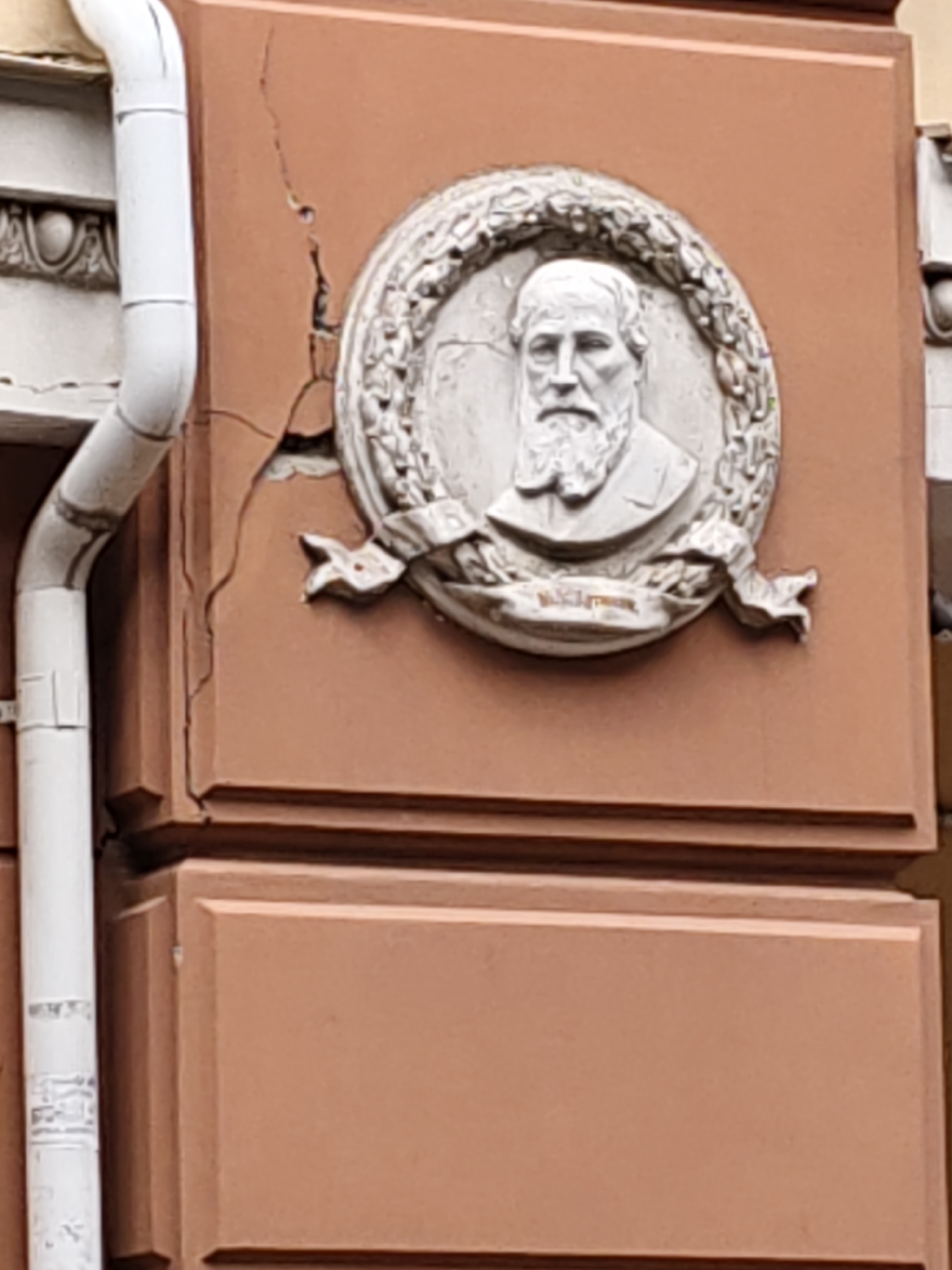 The image shows a bas-relief plaque depicting a bearded man within a wreath, mounted on the exterior wall of a building. The plaque is circular and features the man's bust, facing forward, with a formal suit and a beard. A ribbon with inscription is visible below the bust. The building wall is reddish-brown and shows visible cracks and peeling plaster around the plaque. A white downspout runs vertically along the left side of the building. The architectural style suggests an older building, possibly historic. The scene is captured from a slightly low angle, looking up at the plaque and the building facade.
The bas-relief is located on the building at **ulitsa Bolshaya Sadovaya, 15, Rostov-on-Don, Russia**. This building is known as the former **Azov-Don Commercial Bank building**, and the bas-relief commemorates **Sergey Kirsanov-Borovikov**, a prominent Rostov entrepreneur and founder of the bank.