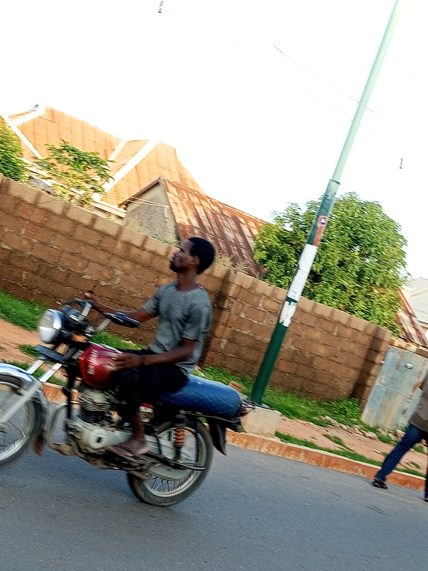 A man is riding a motorcycle on a paved road in Gombe, Nigeria. The man is wearing a grey t-shirt and dark pants, and he is looking to his right. The motorcycle is red and silver. In the background, there is a brick wall and a few houses with corrugated metal roofs. There is also a tall green pole with white signs on it. The sky is overcast. The overall scene suggests a typical street scene in Gombe, Nigeria.