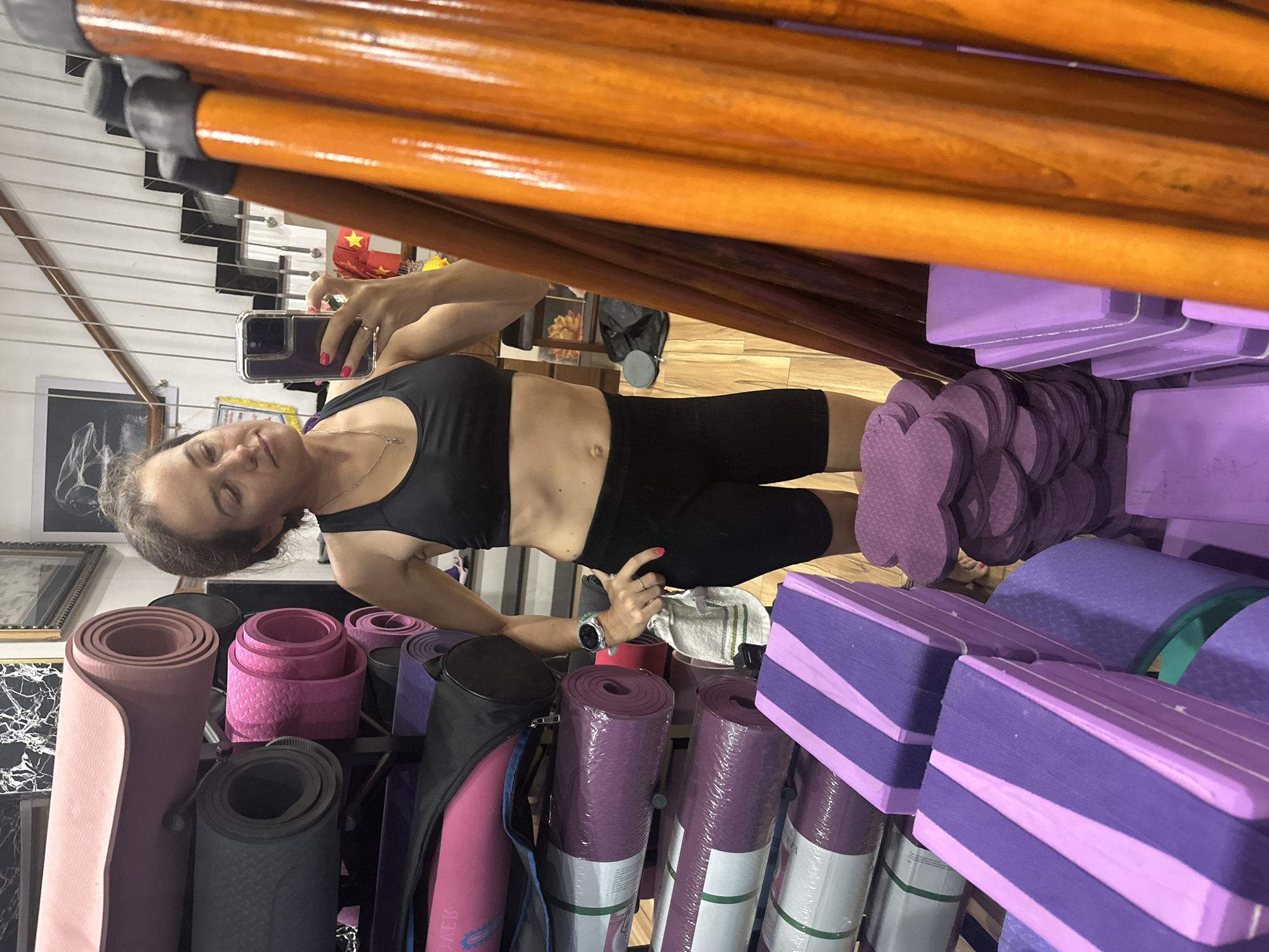 The image shows a woman in a black sports bra and black shorts taking a selfie in what appears to be a yoga studio or store. She is standing next to shelves filled with yoga mats, blocks, and other equipment. The shelves are predominantly filled with purple and pink items. The location may be in Bac Nha Trang Ward, Vietnam.