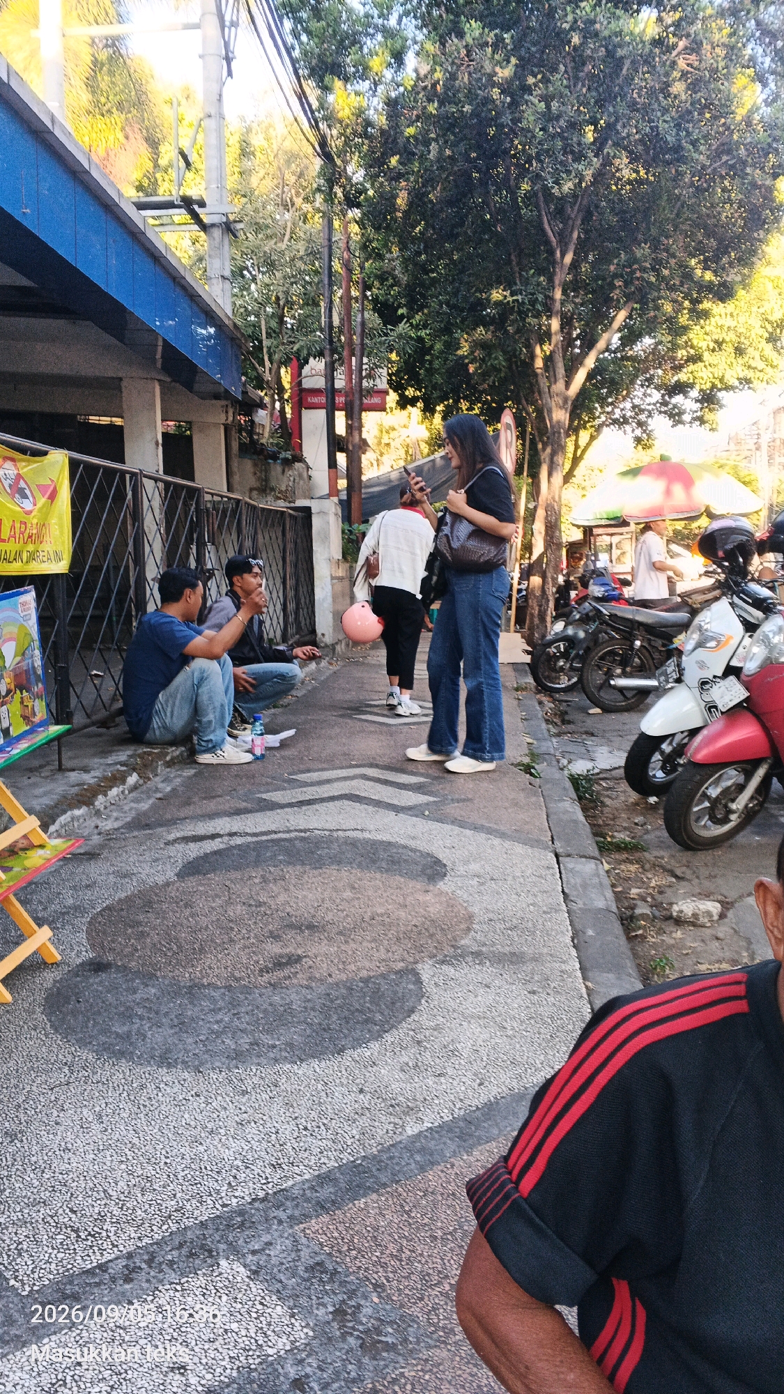 The image captures a street scene in Malang, Indonesia, likely on Jalan Merdeka Barat, given the context of the historical building and the presence of various vendors and pedestrian activity.
In the foreground, a person is partially visible from the shoulder down, wearing a black shirt with red stripes on the sleeves. The ground in front of them is a paved walkway with circular patterns.
Further back, on the sidewalk, three individuals are seated on the ground near a metal fence. The person on the left is wearing a blue t-shirt and jeans, holding a bottle. The person in the middle is wearing a black jacket and jeans, with a black baseball cap. The person on the right is partially obscured but appears to be wearing dark clothing.
To the right of the seated individuals, a woman stands, holding a smartphone in her right hand and looking down at it. She is wearing a black top and blue flared jeans. Beside her, a pink inflatable ball is on the ground. Another person in a white jacket and black pants stands further down the sidewalk.
In the background, several motorcycles are parked along the street. A few vendors are visible, one standing behind a stall with a red and green umbrella. The environment suggests a bustling urban area with a mix of commercial activity and public spaces. Overhead wires and trees are also prominent features of the scene.