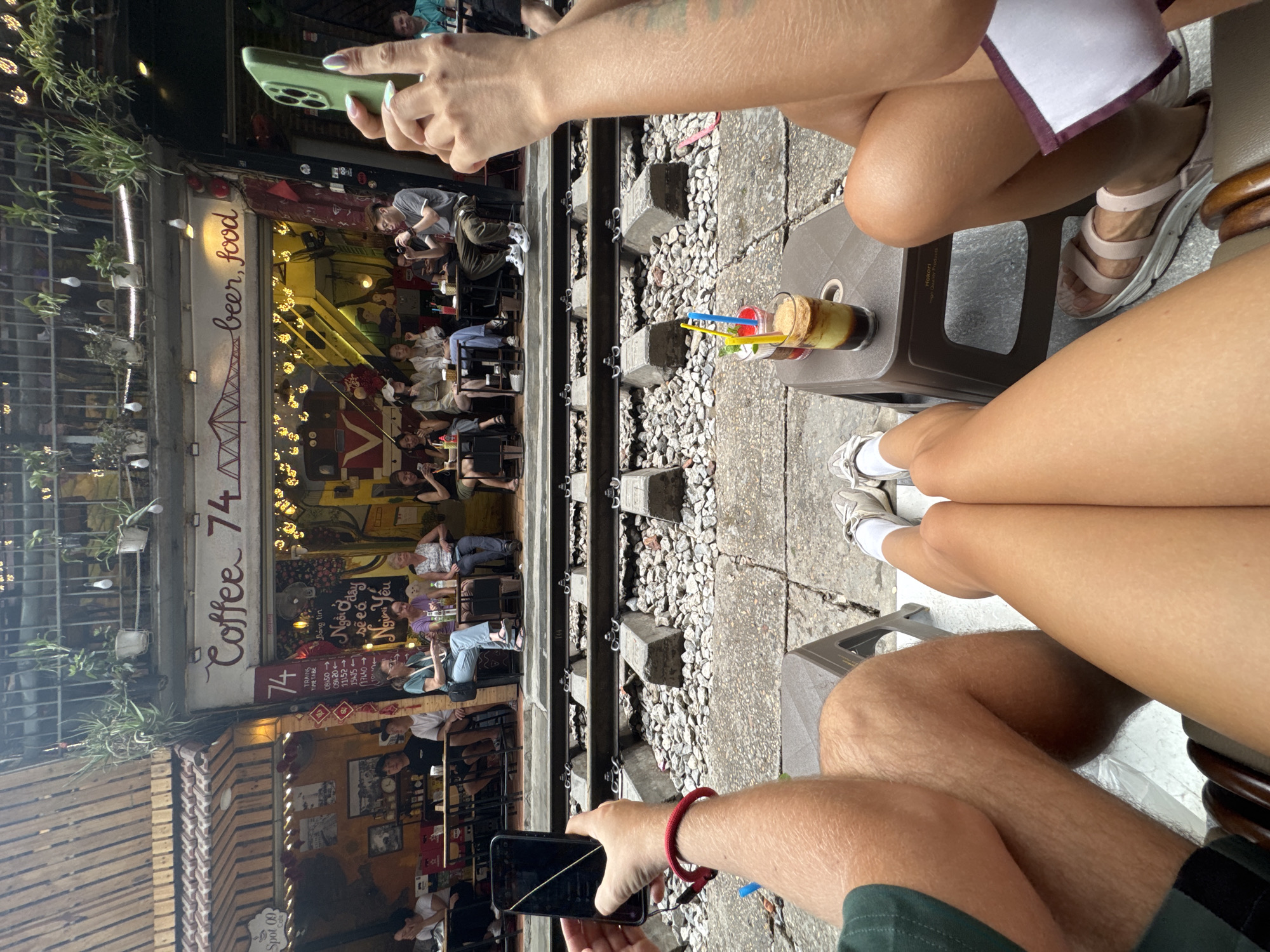 The image captures a lively scene at "Coffee 74" in Hà Nội, Vietnam, positioned alongside operational railway tracks. Three individuals, presumably tourists, are seated with their legs visible, and two are holding smartphones to capture the moment. One is using a green iPhone, and another is using a black smartphone. Their attention is focused on the environment around them, as evidenced by the phones pointed towards the café and tracks. There are two glasses filled with beverages and straws on a small table. "Coffee 74" is adorned with lights, and it appears to be occupied by numerous patrons seated inside. A sign reading "beer, food" is visible, as is a train timetable.