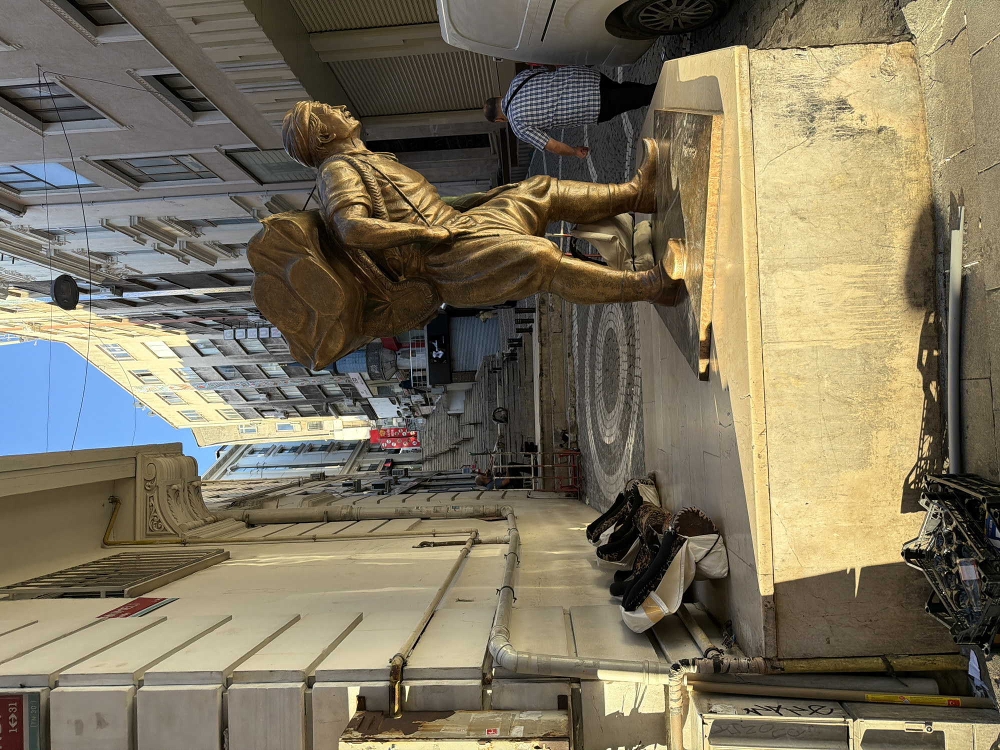 This is a medium shot of a bronze statue of a man carrying a large sack on his back, located on a marble pedestal on a cobblestone street in Istanbul, Turkey. The statue depicts the man bent over, with his head bowed and looking to his right. He is wearing traditional clothing, including a turban, a tunic, and baggy pants tucked into boots. The sack appears to be made of coarse material.
In the background, there is a steep staircase leading up a narrow street lined with multi-story buildings. Several people are visible on the stairs and in the street. A man in a blue and white plaid shirt walks away from the camera to the right of the statue, near a white van. To the left of the statue, near the wall of a building, are several large, rolled-up rugs or textiles, and some stacked black plastic crates. The street is illuminated by sunlight, casting shadows. The buildings are a mix of architectural styles, with some featuring ornate details and others being more modern. The overall scene suggests a busy urban environment with a historical feel.
