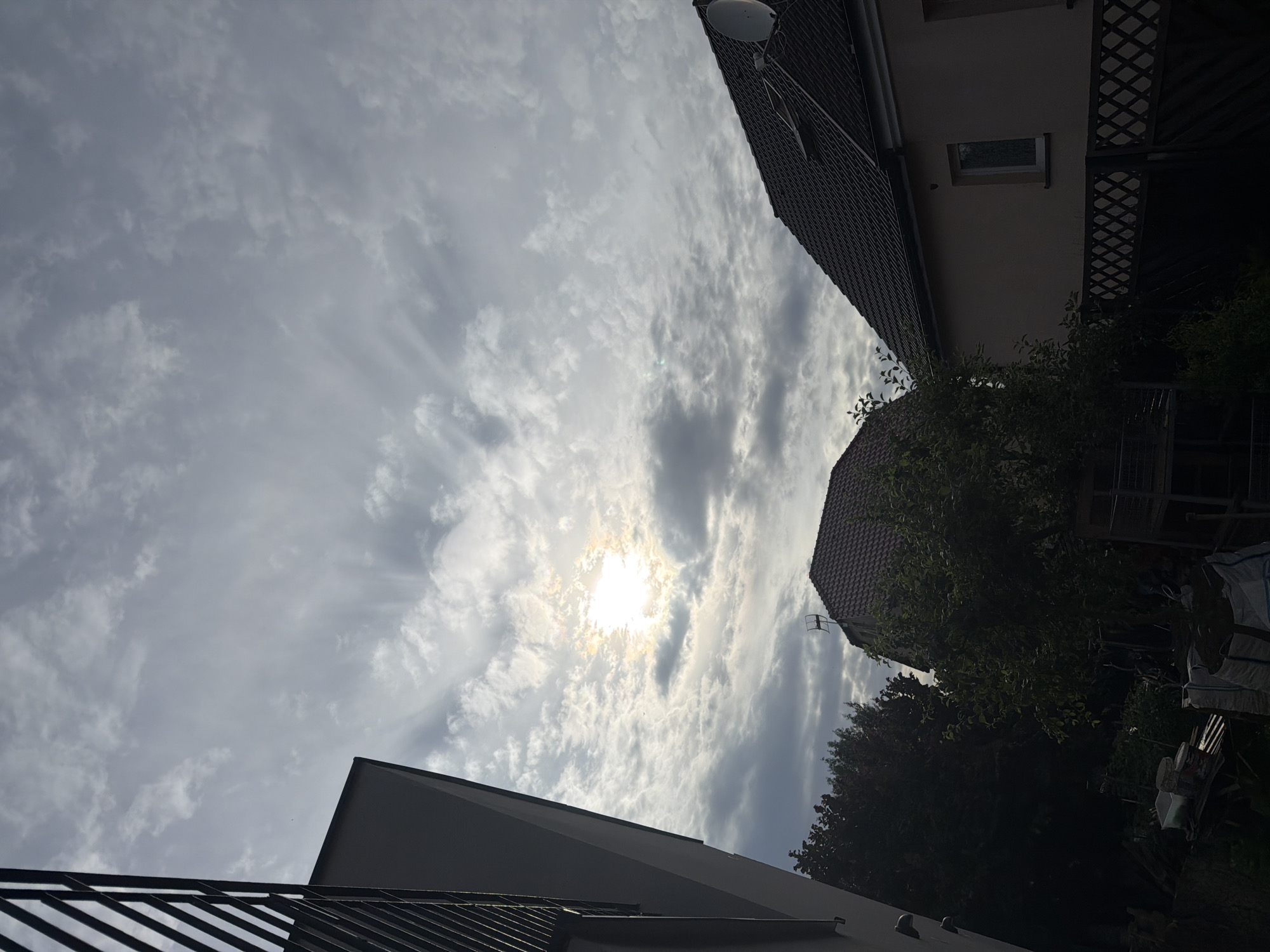 This media file captures an outdoor scene, likely a residential area in Szczecin, Poland. The viewpoint is looking upwards towards a partly cloudy sky where the sun is visible, creating a bright, somewhat diffused light. Streaks of clouds radiate outwards from the sun, suggesting a dynamic atmospheric condition.
In the foreground, parts of building rooftops are visible. On the left, a dark, possibly metal roof with parallel lines is prominent. To the right and further back, the dark, tiled roof of another building is seen. Between these structures, lush green trees contribute to the natural element of the scene. The sky, with its scattered clouds and sunshine, indicates it is daytime, likely late morning or afternoon, with moderate weather conditions. The overall mood is serene, with the natural light playing a significant role. There are no visible people or text in the image. The presence of buildings and trees suggests an urban or suburban environment.