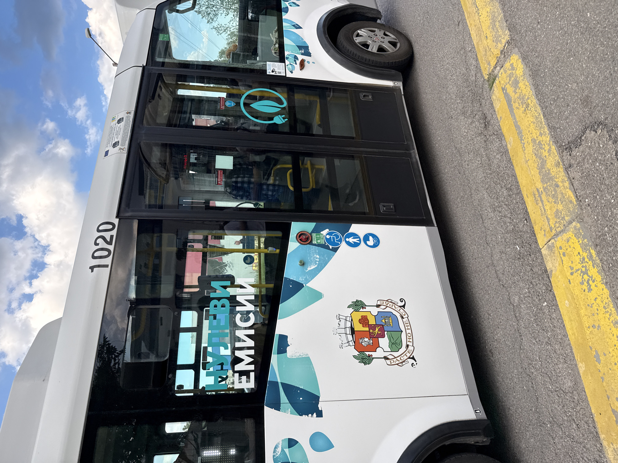 A white electric bus with the number 1020 is parked on a street with a yellow curb. The bus has graphics related to electric vehicles, the phrase "Zero Emissions" (in Bulgarian), and the coat of arms of Sofia, Bulgaria. Based on the coat of arms and language used on the bus, it is located in Sofia, Bulgaria.