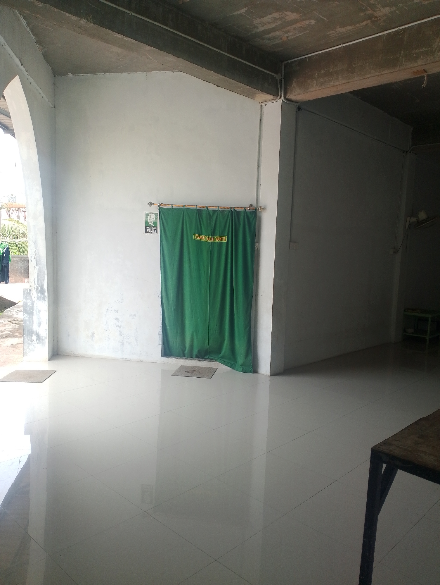 The image shows an interior view of what appears to be a public restroom or changing area, possibly within a mosque or community center in Batam, Indonesia. A prominent feature is a large, dark green curtain hanging from a bamboo rod, concealing an entrance. Above the curtain, a sign with green lettering on a white background is visible, reading "TEMPAT WUDHU WANITA," which translates to "Women's ablution place." To the left of the curtain, there is another smaller sign with a picture and text, also in green. The floor is tiled with large, light-colored square tiles that reflect light. Two dark-colored mats are placed on the floor, one near the entrance to the curtained area and another further to the left near an open archway. Through the archway, in the background, two women wearing headscarves are visible. The walls are painted white and appear somewhat textured. The ceiling is a concrete structure with visible beams and conduits for electrical wiring. In the far right of the image, a dark-colored table with a rustic appearance is partially visible, and beyond it, a dimly lit area with a small green shelf unit can be seen. The overall impression is one of a utilitarian and functional space.