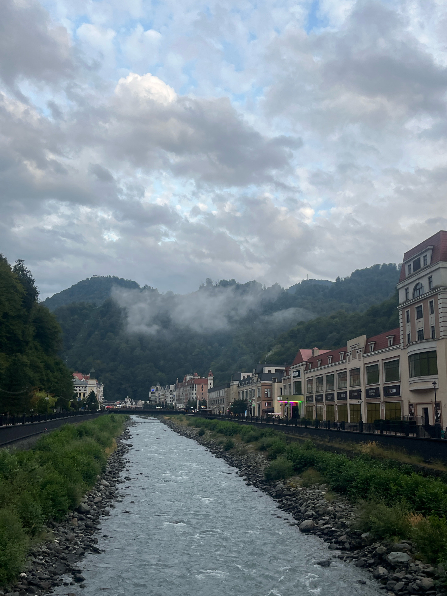 The image depicts a view along a river in the city of Estosadok, Russia, with a mountain range in the background. The river flows from the distance towards the foreground, its surface showing ripples and currents. On the right bank of the river, a row of buildings with distinct architectural styles, including red tiled roofs and light-colored facades, is visible. Some of these buildings appear to be shops or commercial establishments, with visible signage. A paved walkway runs alongside the buildings. On the left bank, dense green foliage covers the slope leading up to a forested mountainside. The mountains in the background are shrouded in clouds and mist, adding a dramatic and atmospheric quality to the scene. The sky above is overcast with gray clouds, suggesting a possibly cool or damp day. There are no people clearly visible in the image. The overall impression is of a picturesque mountain resort town.