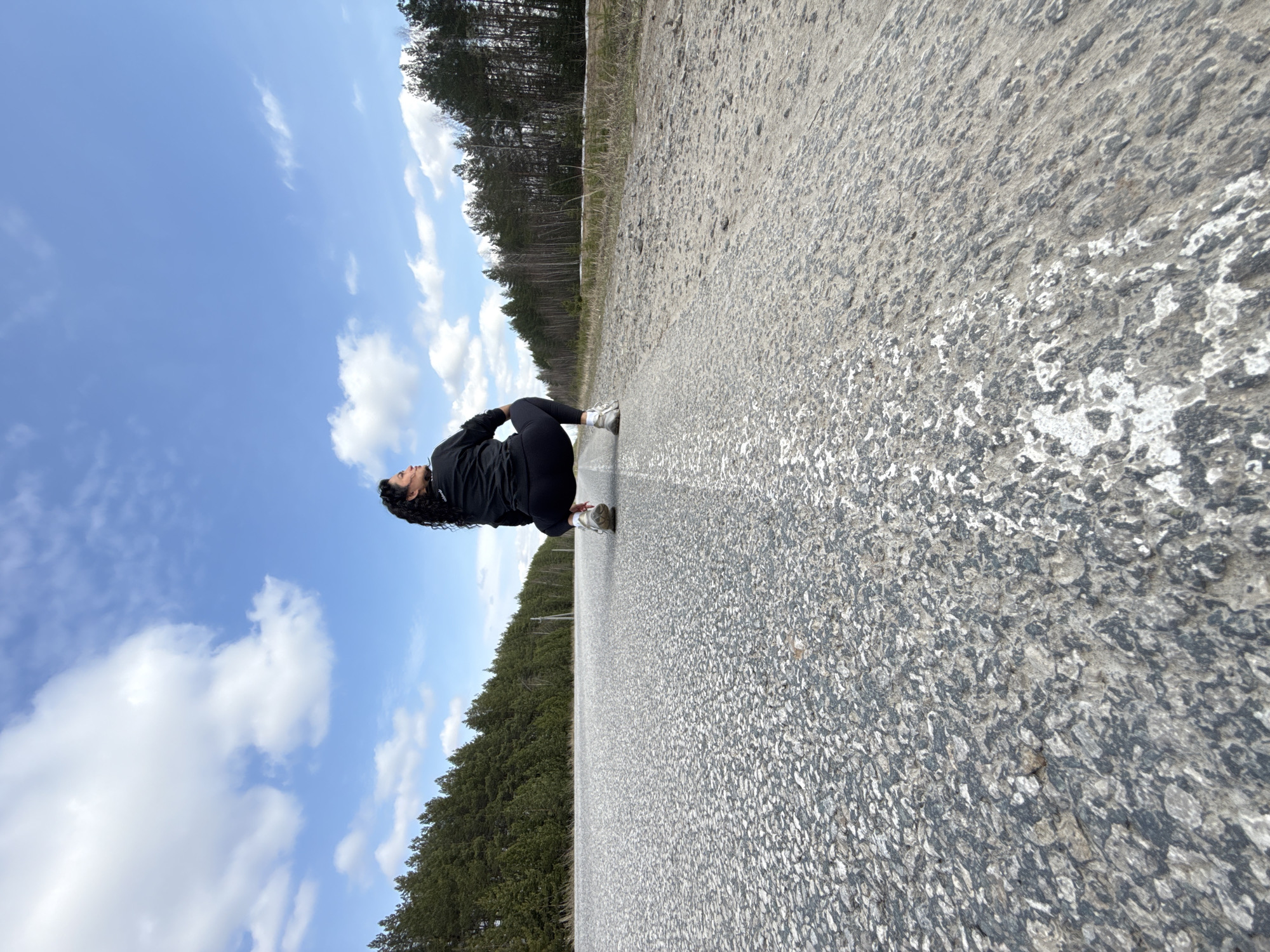 The image, when correctly oriented, depicts a person crouching on the edge of a long, straight paved road, flanked by a dense forest under a blue sky. The subject, a person with long, dark curly hair, is wearing a black long-sleeved top, black leggings, and white sneakers. They are in a low squatting or crouched position on the road's surface, looking towards the left side of the frame with their arms bent.
The setting is a quiet, possibly rural or remote, area. The road surface is grey and textured, appearing somewhat worn, with indistinct lighter markings visible on the right side. On both sides of the road, a thick forest of tall, dark green coniferous trees stretches into the distance, suggesting a natural, unpopulated landscape. Above, the sky is a bright blue, dotted with scattered white, fluffy clouds, indicating clear and pleasant weather. The time of day appears to be daytime, likely morning or afternoon, given the natural light. The overall mood is calm and reflective. The scene is located in Maksimovo, Russia. No visible text is present in the image.
