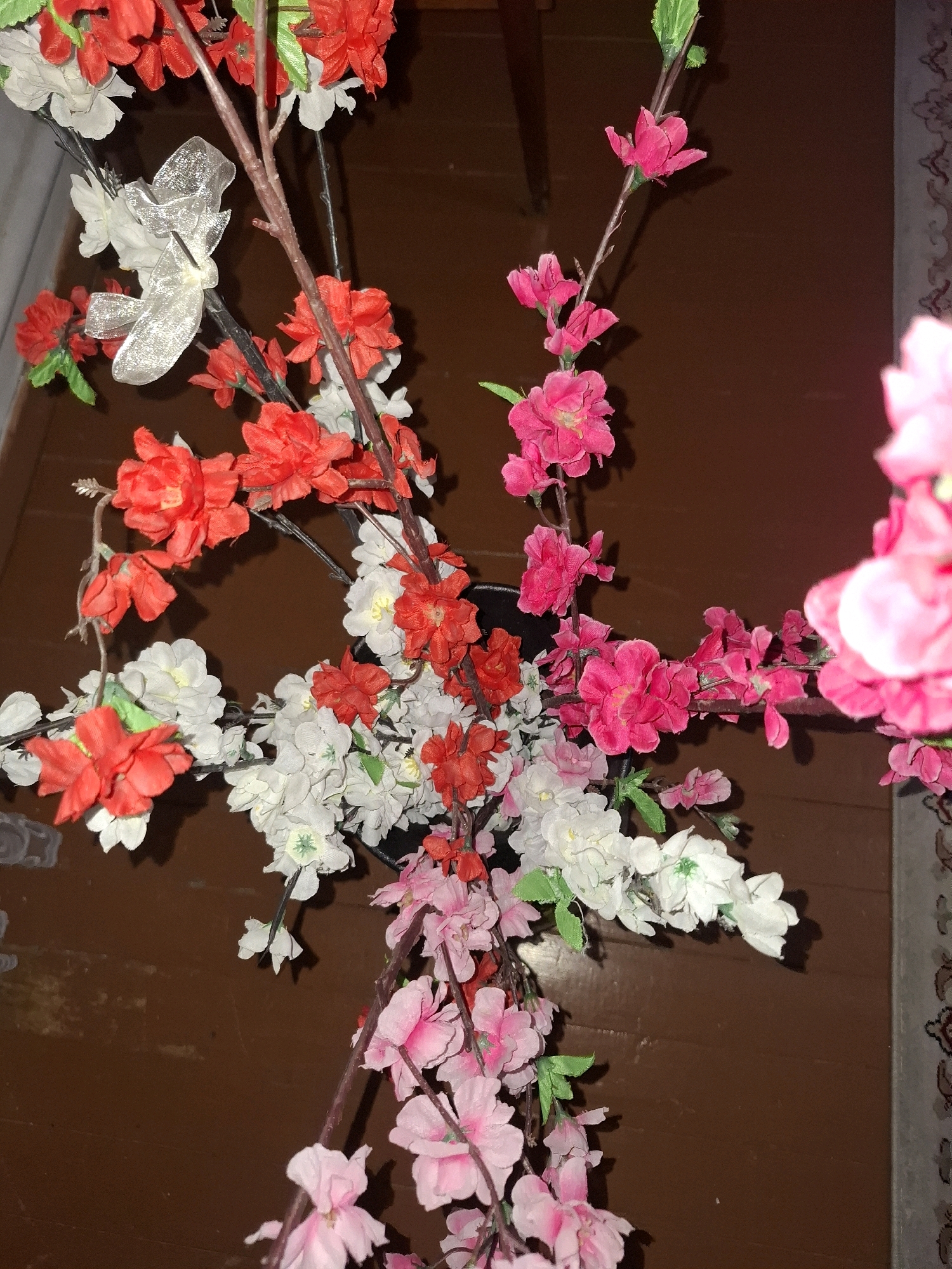 A close-up, vertical shot showcases an artificial floral arrangement, predominantly featuring pink, white, and red blossoms. The flowers are attached to thin, brown branches, some adorned with small green leaves. A silvery ribbon bow is visible on one of the branches. The arrangement is set against a dark brown background, likely a wooden surface. To the right edge, a portion of a patterned textile is visible. The lighting in the image is dim, with some highlights on the flowers and ribbon, and a soft shadow cast on the background. There are no people or animals present. The scene appears to be indoors, possibly a home or event space, and does not provide any specific visual cues to pinpoint a location within Almaty, Kazakhstan.