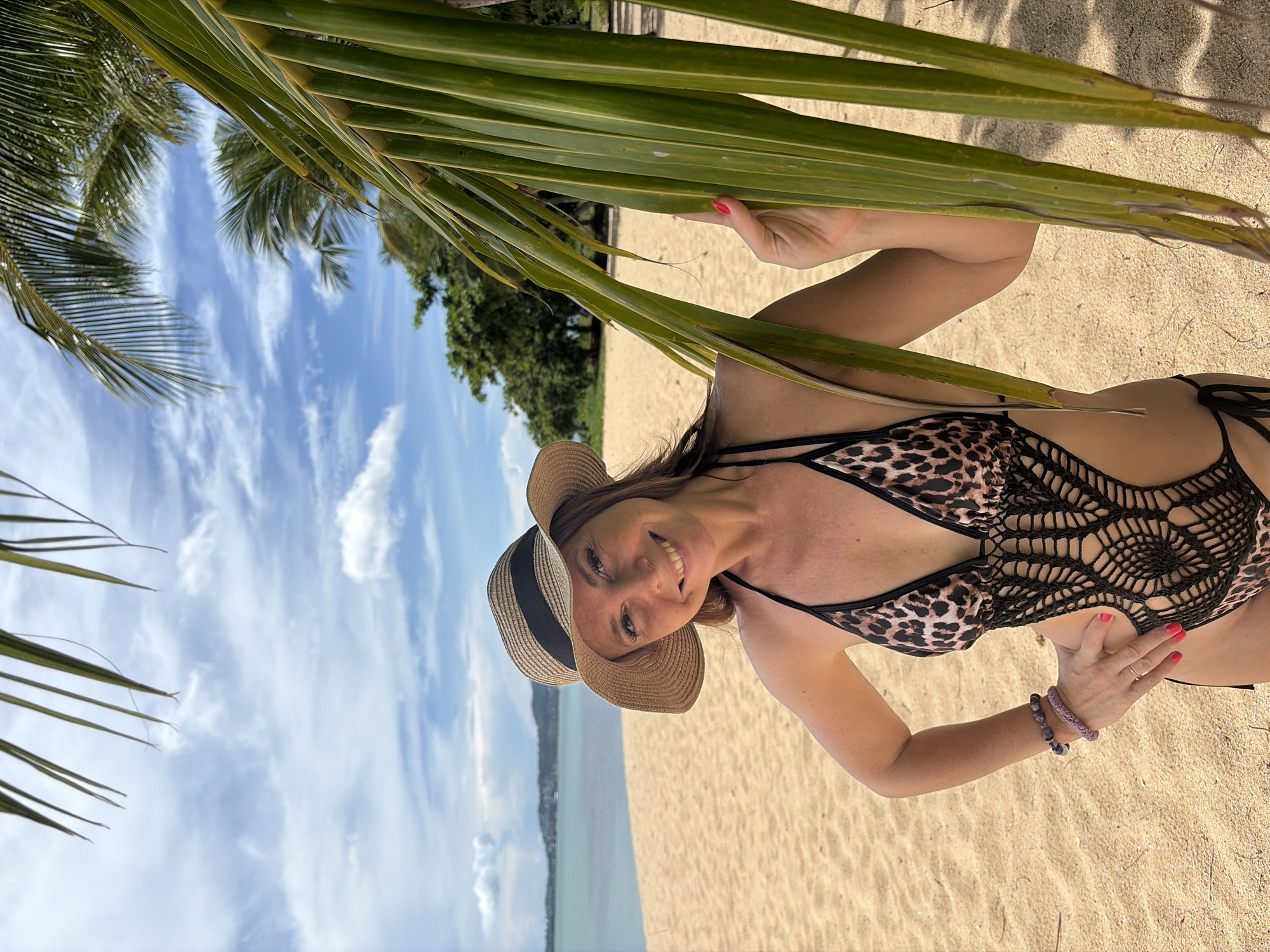 A smiling woman wearing a leopard print one-piece swimsuit with crochet detailing and a straw hat stands on a sandy beach. She is partially obscured by palm fronds. In the background, the turquoise ocean meets the horizon, with a distant shoreline and a cloudy blue sky. The image was taken in Baan Bo Phut, Thailand.