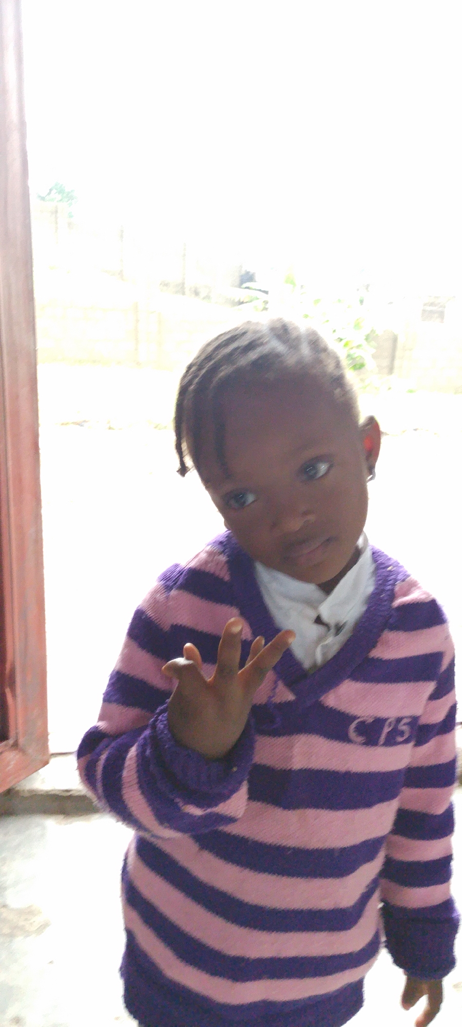A young child, presumably a girl, is standing indoors near a window in Lokoja, Nigeria. She is wearing a pink and purple striped sweater with a white collared shirt underneath. Her hair is braided. She is holding up her hand, seemingly making a gesture. The background shows a blurred outdoor scene, possibly a yard or street. The light suggests it is daytime.