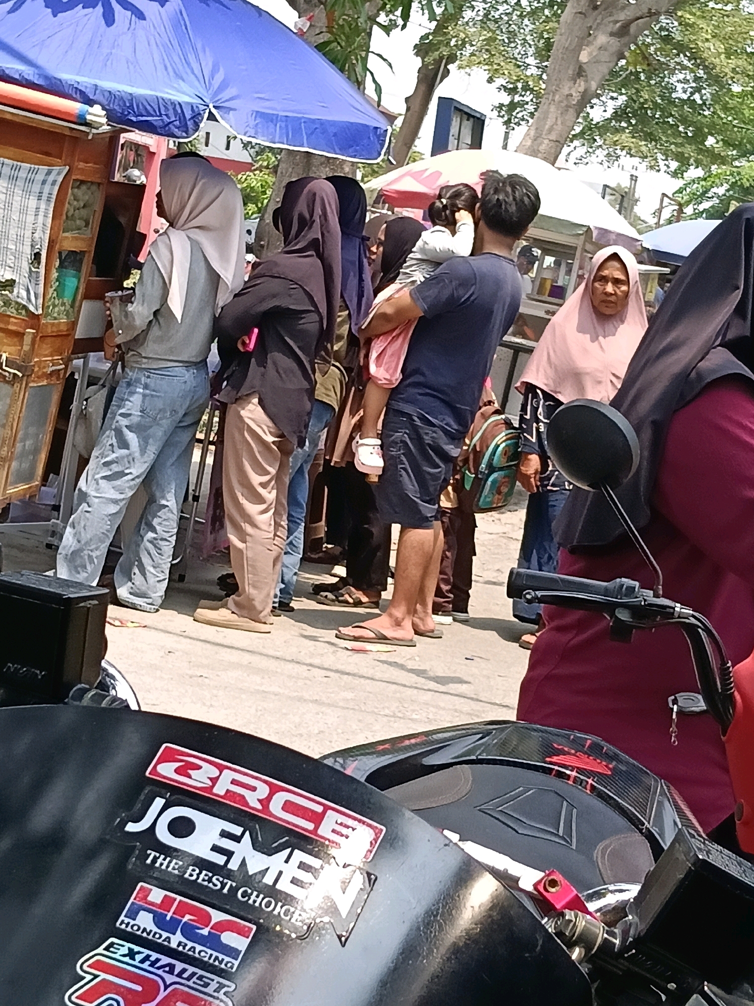This is a medium-shot, eye-level photograph taken outdoors in the city of Belimbing, Indonesia. The scene depicts a bustling street with several people and market stalls. In the foreground, a black motorcycle is partially visible, adorned with several stickers, including "ORCE," "JOEMEN," "THE BEST CHOICE," "HRC HONDA RACING," and "EXHAUST." The motorcycle's handlebars and side mirror are also visible.
Behind the motorcycle, a group of people are seen standing. A woman wearing a light-colored hijab and a grey top is looking to her left, holding what appears to be a cup or container. Next to her, another woman in a black hijab and dark clothing is looking forward. A man in a dark blue t-shirt and dark shorts is holding a young child in a pink outfit. Further back, a woman in a pink hijab is looking towards the camera. Other individuals are visible in the background, including a person in a blue t-shirt and another in dark trousers and a yellow top, possibly a vendor.
Market stalls are set up, with one stall featuring a blue tarp overhead and wooden structures displaying goods. Another stall has a white and pink umbrella. The environment appears to be a sunny day, with trees and foliage visible in the background, suggesting a tropical or subtropical climate. The ground is paved with concrete. The overall impression is of a busy marketplace or street scene in Indonesia.