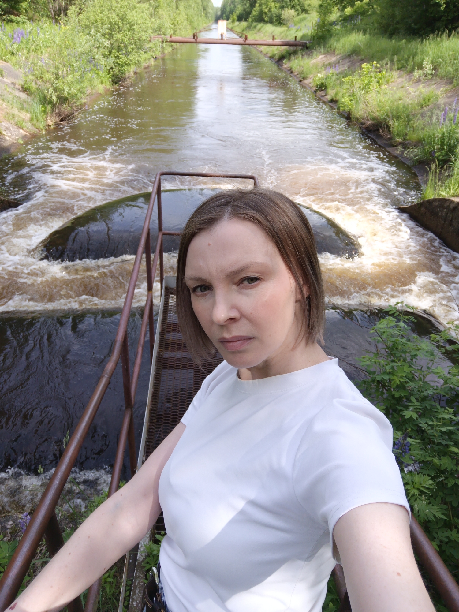 A woman is captured in a medium-shot selfie, positioned on a rusty brown metal grate walkway with railings to her left, overlooking a canal. She appears to be in her 30s or 40s, with short, light brown hair and a fair complexion. She wears a plain white short-sleeved t-shirt and gazes directly at the camera with a neutral, slightly serious expression. Part of her left arm is visible, suggesting she is holding the camera.
The scene behind her features a canal with swiftly flowing, murky brown water. The water churns into white foam as it cascades over two dark-colored spillways or weirs, creating small, turbulent waterfalls. The banks of the canal are lush with green vegetation, including tall grasses, bushes, and scattered purple flowers, likely lupines. The banks appear to be reinforced, possibly with concrete structures.
In the mid-distance, a long, reddish-brown pipe or aqueduct spans horizontally across the canal. The waterway continues into the background, flanked by dense green trees. The image is taken during daylight hours, indicated by bright illumination, and the weather appears clear. There is no visible text in the image. The setting suggests an outdoor, possibly rural or industrial, environment involving water management infrastructure in Chernushka, Russia.