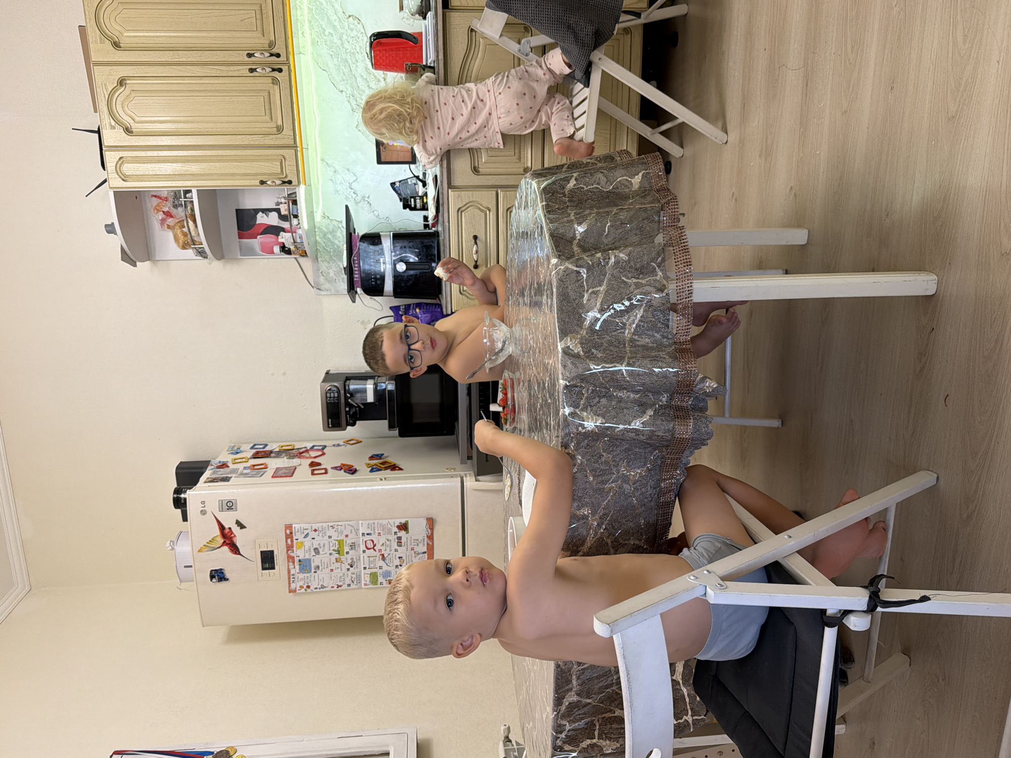 This media file captures a lively scene in a home kitchen, likely in Sochi, Russia, during daytime. The image shows three young children engaged in different activities around a kitchen table and counter.
In the foreground, a shirtless young boy with short blonde hair and grey shorts sits at a white folding chair by a table, looking slightly to the side. The table is covered with a clear plastic tablecloth over a patterned fabric. Beside him, another shirtless boy with short brown hair and glasses is also seated at the table, looking directly at the camera. He holds a spoon and has a bowl, possibly containing strawberries, in front of him. Both boys appear to be eating a meal or snack.
In the background, a toddler with blonde curly hair, dressed in pink polka-dot pajamas, stands on a white folding chair and reaches towards the kitchen counter. This suggests curiosity or an attempt to grab something from the counter.
The kitchen features light-colored wooden cabinets above and below a greenish-marbled countertop. Various kitchen appliances are visible on the counter, including a black air fryer, a coffee maker, and a red toaster or kettle. An open upper cabinet reveals a bag of bread. A white LG refrigerator is adorned with numerous magnets, including a hummingbird, and several papers or charts with visible text, including "МАЙ" (May) in Cyrillillic script, suggesting a calendar or schedule. The floor is covered with wood-look laminate. The overall impression is a casual, lived-in family kitchen environment.