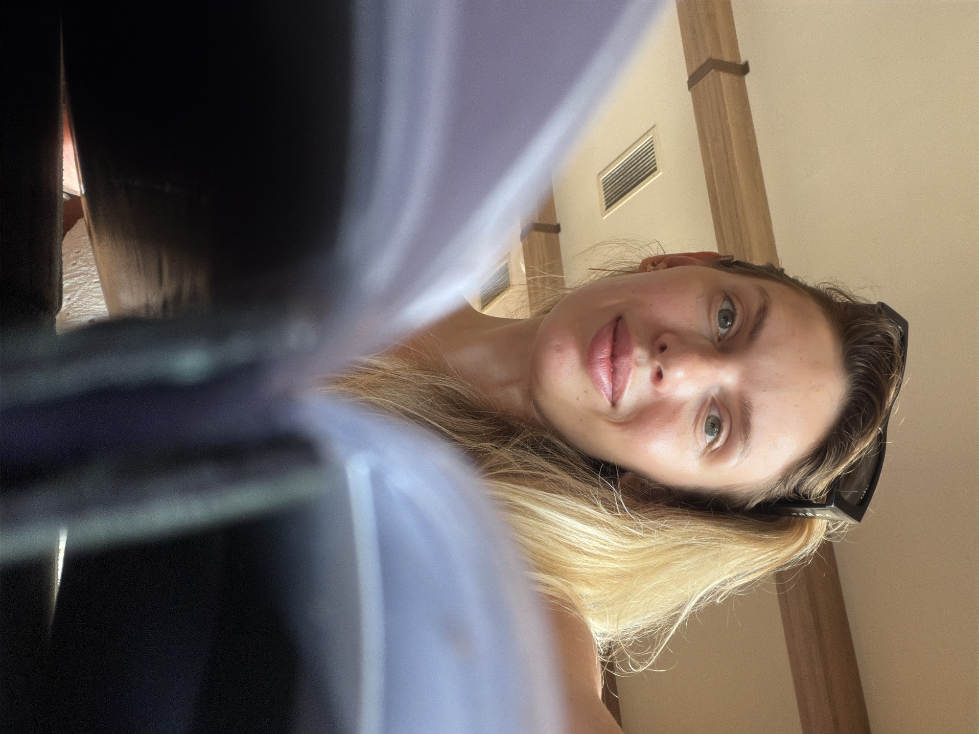 A woman with blonde hair pulled back and sunglasses perched on her head is looking into the camera from behind an open book. She is indoors, with a ceiling fan vent and wooden beams visible in the background. The lighting suggests it is daytime. She appears to be in a relaxed setting, possibly a cafe or library. No specific location in Bigor, Montenegro is identifiable from the image.