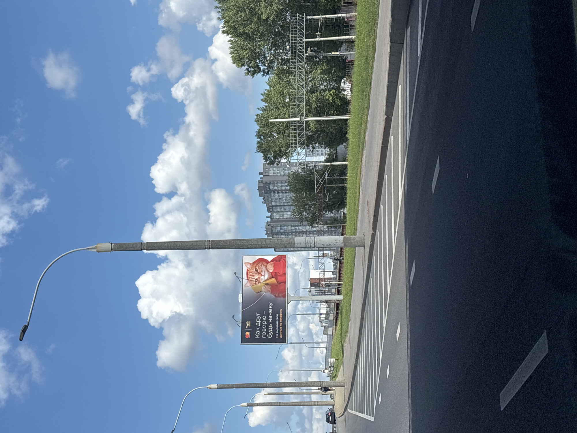 The image shows a street scene in Minsk, Belarus, on a sunny day with a bright blue sky and white, fluffy clouds. In the foreground, a dark asphalt road with white lane markings is visible, leading towards the right side of the frame. To the left of the road, a sidewalk with green grass and a concrete curb runs alongside. Several tall, gray streetlights with curved arms are spaced along the sidewalk.
Dominating the mid-ground is a large billboard featuring a ginger cat wearing a red hoodie and holding a yellow smartphone to its ear. The billboard displays Russian text, which translates to "As your friend says - be on guard. Don't give away your phone data." This suggests the billboard is an advertisement for a financial or security service.
Behind the billboard, a row of trees with lush green foliage lines the horizon. Further in the background, several tall, apartment buildings are visible, characteristic of urban residential areas. Railway tracks and electrical lines are also present, indicating proximity to transportation infrastructure. A few cars and a person walking are visible on the road in the distance. The overall impression is a typical urban environment on a clear, pleasant day.