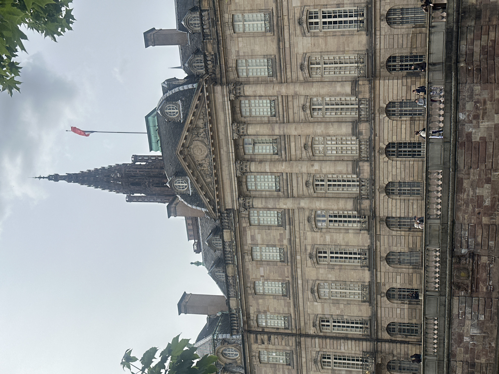 The image captures a view of the Rohan Palace in Strasbourg, France, with the Strasbourg Cathedral spire visible in the background. The Rohan Palace, a large baroque building, features a stone facade with multiple rows of windows and decorative architectural elements. A bridge or walkway made of stone runs in the foreground, with several people strolling along it. The sky is overcast, and some tree branches with leaves are visible in the upper left and right corners.