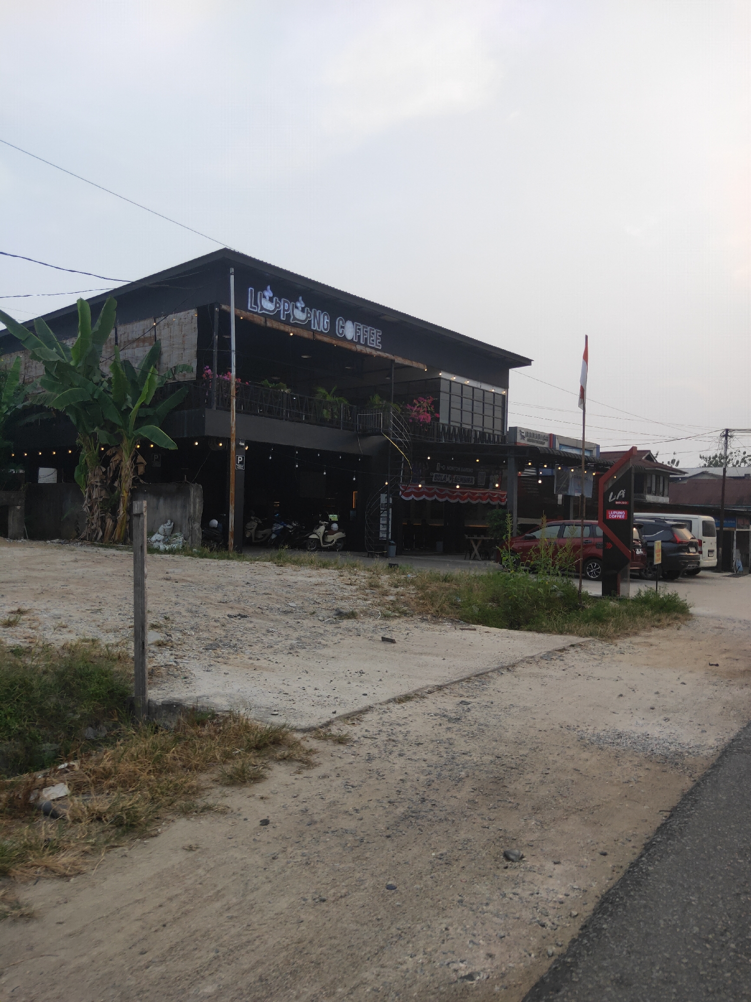 The image depicts a two-story building, likely a coffee shop, with a sign that reads "LUPUNG COFFEE" in illuminated lettering. The building has a dark, modern facade with a balcony on the second floor, adorned with potted plants and decorative flowers. Several motorcycles are parked in the open-air ground level of the building. To the right of the coffee shop, a red car and a dark SUV are parked. A red and white Indonesian flag flies on a flagpole near the entrance. The area in front of the building is a dirt and gravel lot, with a paved road visible in the foreground. The scene appears to be in a semi-urban or rural setting, with a clear, overcast sky. The coffee shop is situated along a road in Sintang, Indonesia.