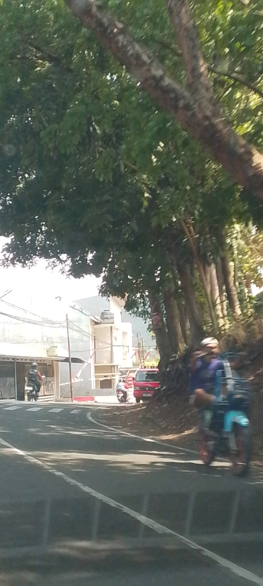 The image depicts a street scene with vehicular traffic and foliage in Susukan, Indonesia. On the left side of the road, a person rides a scooter away from the viewer. In the background, a building complex with a light-colored facade and a water tank on the roof is visible, with power lines running across the front. Further down the road, another scooter is parked near a red vehicle. On the right side of the road, a person is riding a scooter with a blue carrier attached to the rear, moving towards the viewer. The road is asphalt with white lane markings. Lush green trees line the right side of the road, casting shadows on the asphalt. The overall impression is a typical day in a suburban or semi-urban area with moderate activity.