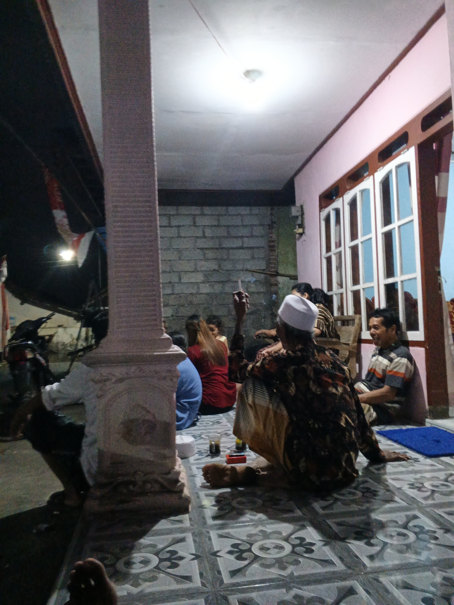 In Singolatren, Indonesia, a group of people are gathered in an outdoor area, likely a veranda or patio, at night. The environment features a patterned tile floor, a pink-painted wall with a large, multi-paned window, and a brick wall. A prominent decorative pillar stands in the foreground, partially obscuring the view of some individuals.
Several people are seated on the floor. One man in the foreground wears a white peci (cap) and a patterned shirt, and appears to be holding a cigarette. Another man, wearing a striped shirt, is seated to the right, looking towards the left and smiling. Further back, a woman in a red top and another person in a blue top are visible, along with others whose faces are not clearly discernible. The lighting suggests artificial illumination from overhead. In the background, a motorcycle is parked near a dark roofed structure. The scene appears to be a casual social gathering.