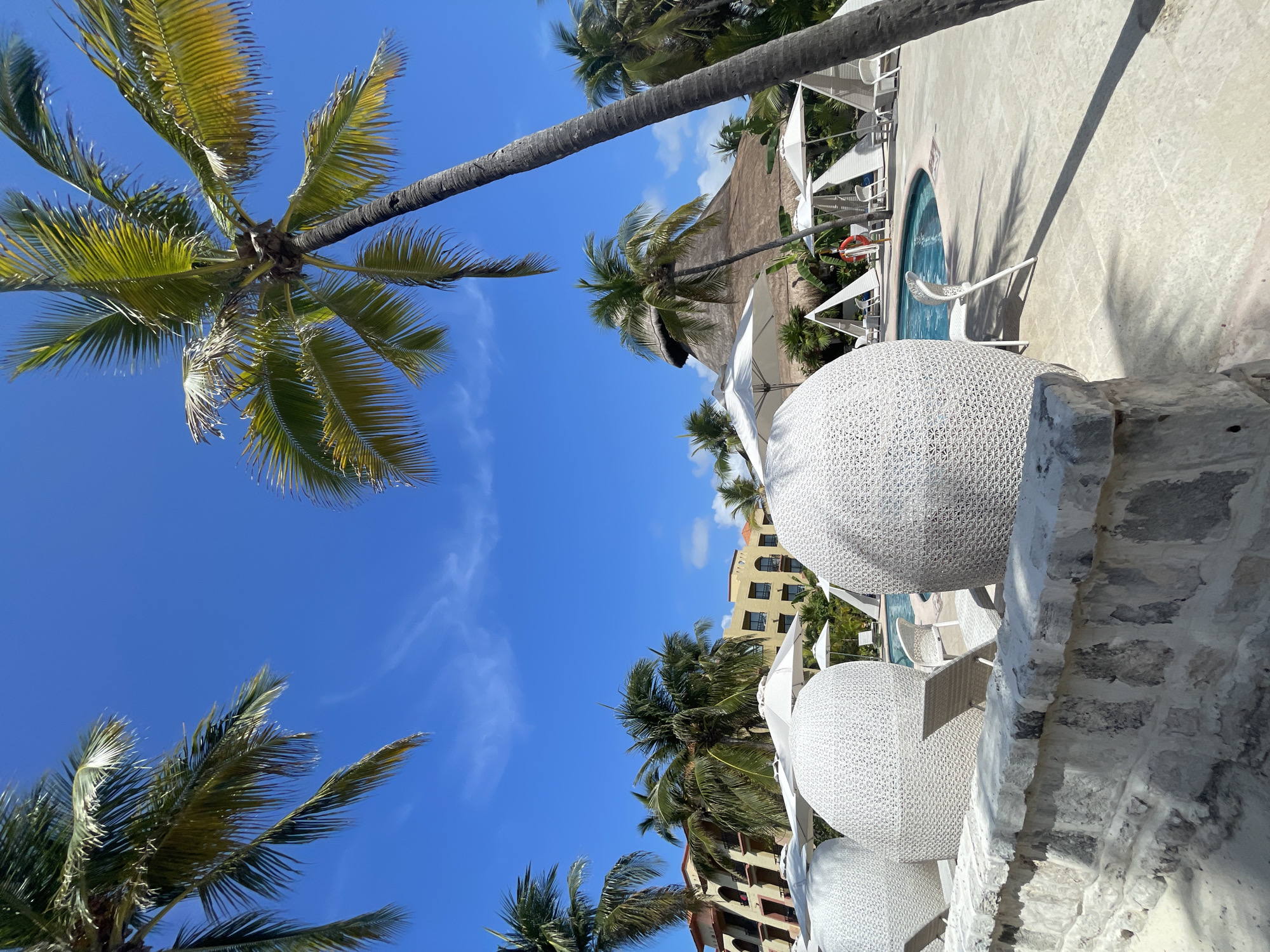 The image shows a sunny tropical scene in Playa del Carmen, Mexico. The view is angled upwards, capturing palm trees against a clear blue sky with scattered clouds. In the foreground, a rough, white stone wall is visible, with white, woven, spherical chairs or decor elements placed on it. In the midground, there's a resort-style area with a small, curved pool, white lounge chairs, and white sun umbrellas or canopies. A thatched roof structure is also present. A multi-story building in light yellow and beige tones can be seen behind the palm trees.